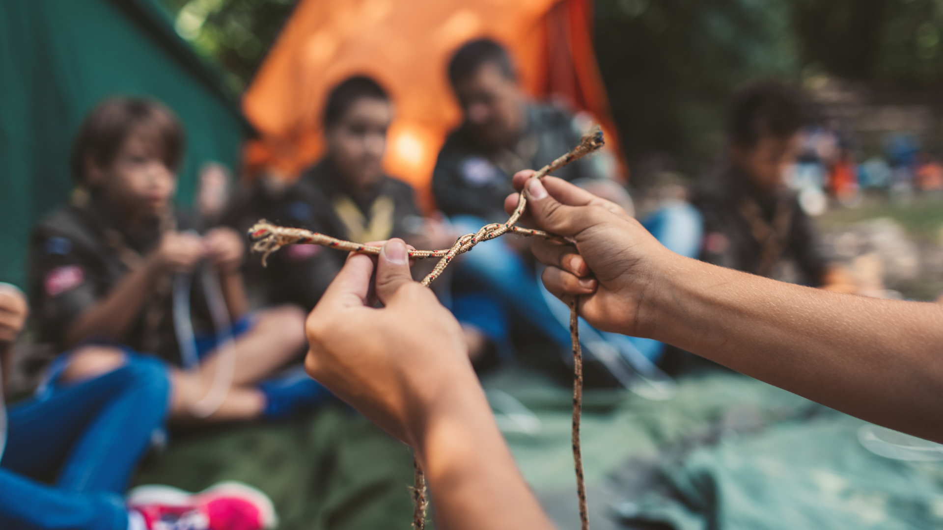 Person tying a knot in a rope with a group of Boy Scouts sitting outdoors in the background.