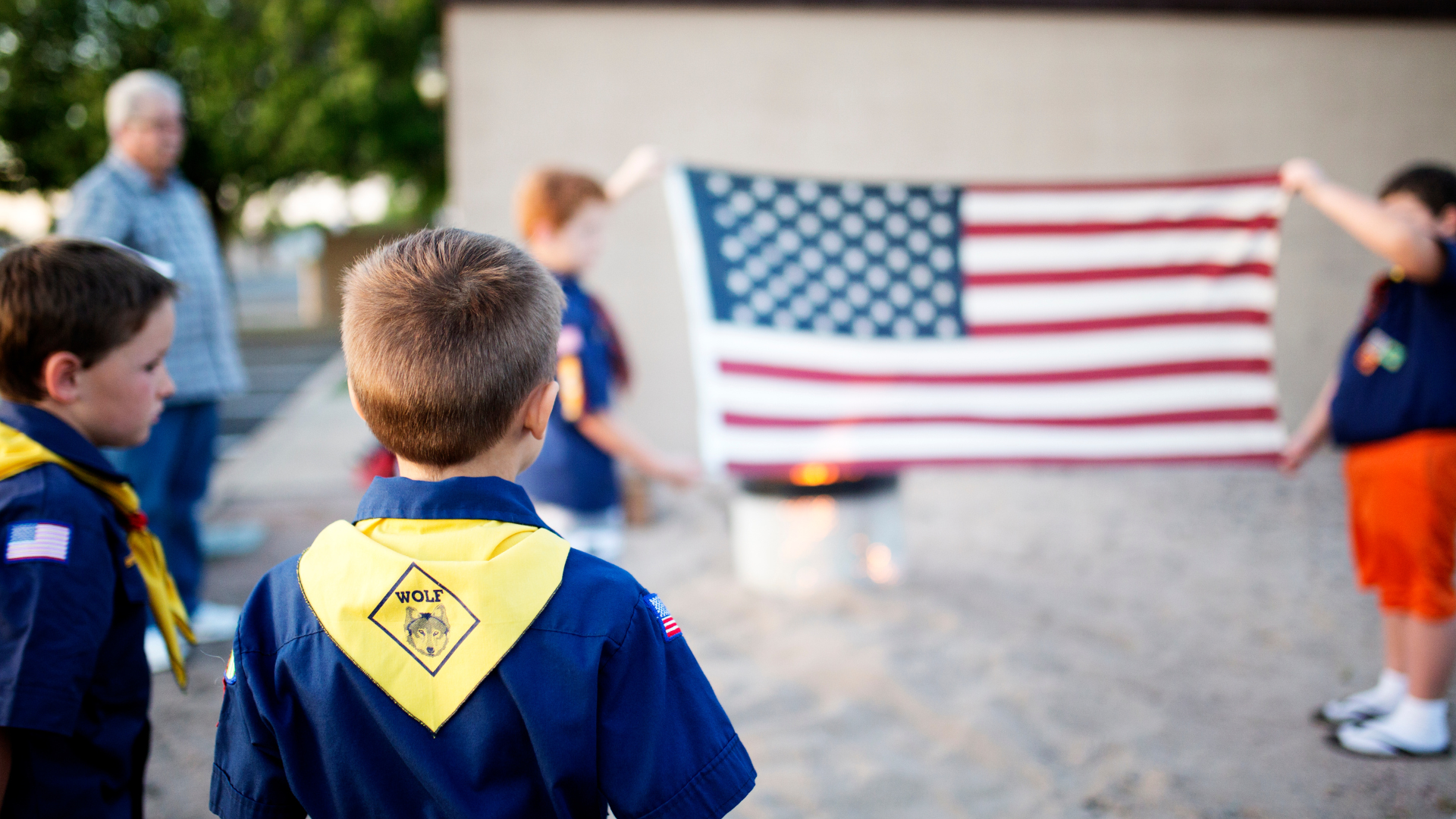 Boy scout in uniform with a yellow neckerchief looking at Boy Scouts holding a large American flag during a ceremony.
