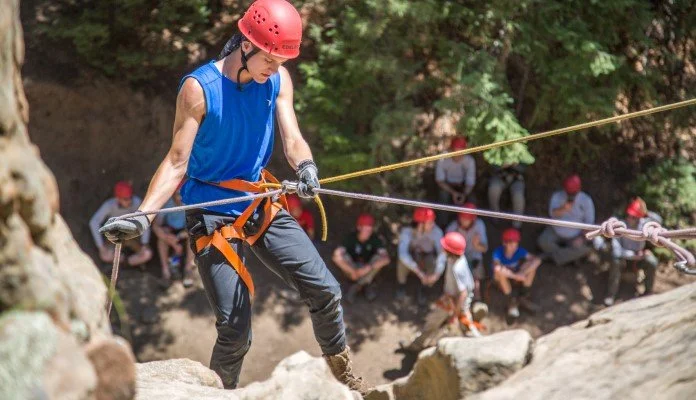 Person in a red helmet rock climbing outdoors with others seated in the background.