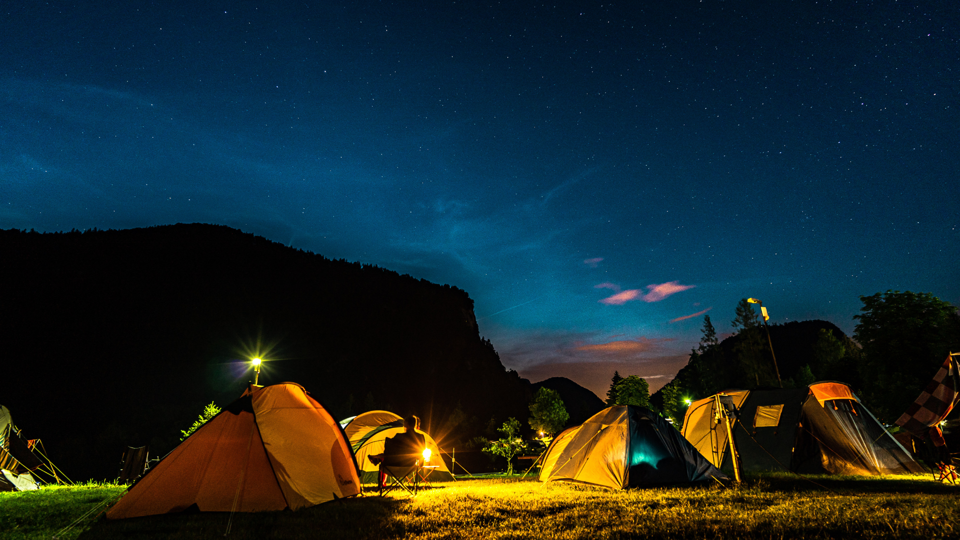 Nighttime scene of tents set up in a grassy area with mountains in the background, under a starry sky. A person is sitting on a chair near a tent, illuminated by camping lights.