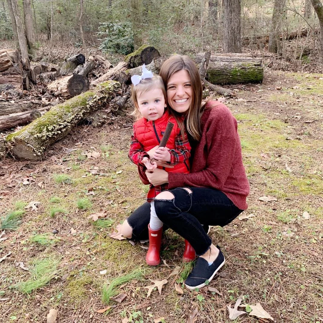 A woman crouching outdoors with a young girl in a wooded area, both smiling. The girl is wearing a red plaid jacket, white bow in her hair, and red rain boots. The woman is wearing a red sweater, ripped black jeans, and black slip-on shoes. There are logs and fallen leaves in the background.