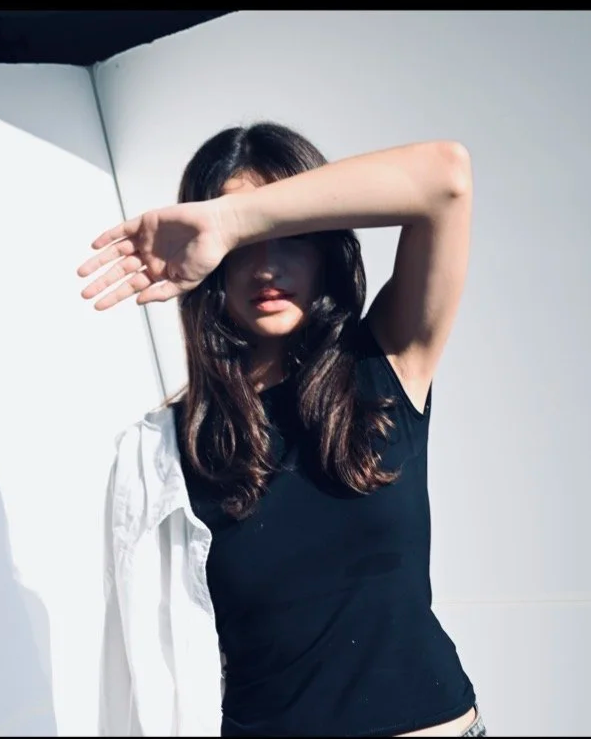 Person in black sleeveless top, partially covering face with arm, standing against a light background with contrast shadows.