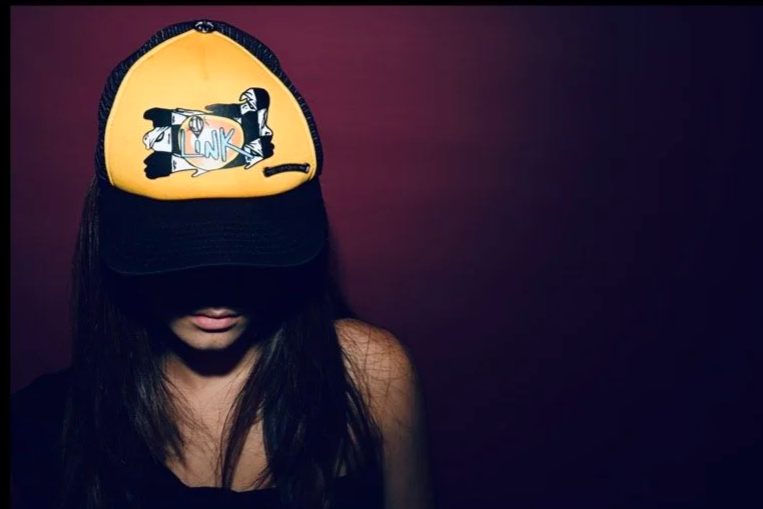 Person wearing a yellow and black cap, shadowed face, dark background, graphic design on cap.