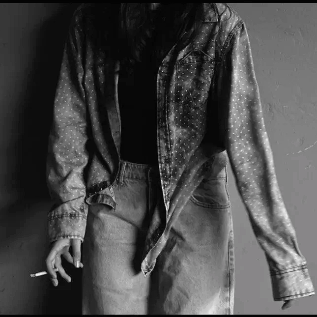 Black and white photo of a person in a denim jacket, holding a cigarette, with only the lower half visible.