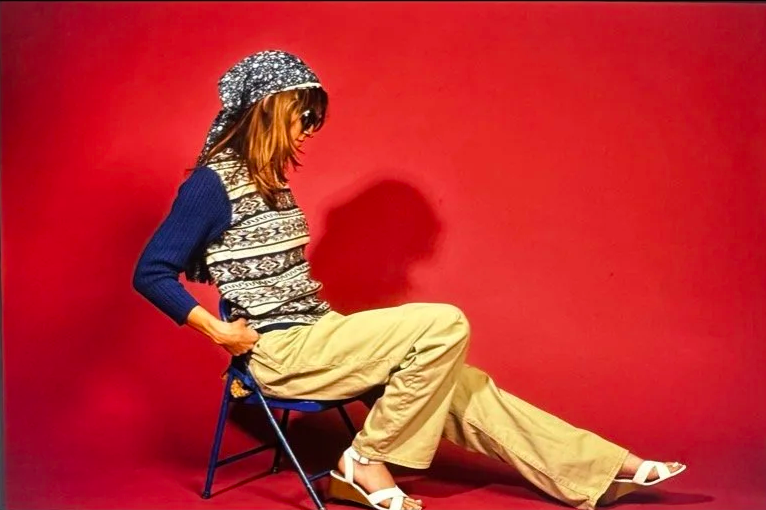 Person wearing patterned top and beanie, sitting on a chair against a red background.