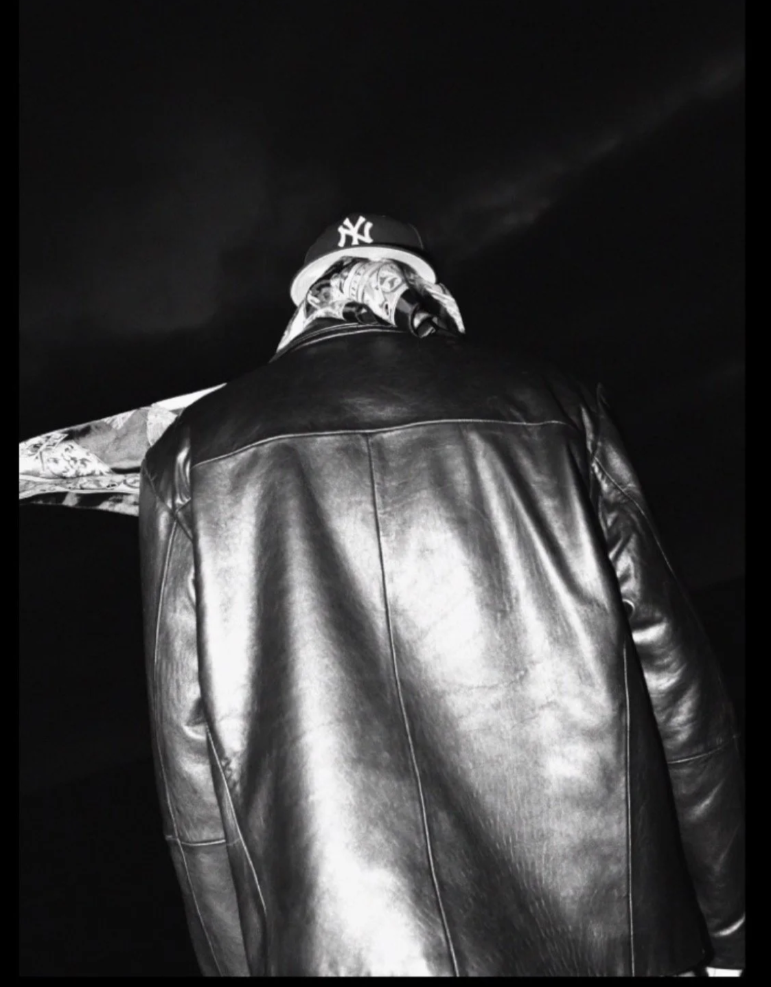 Black and white image of a person wearing a leather jacket and a baseball cap with a bandana tied around their neck. The person is looking away from the camera, against a dark background.