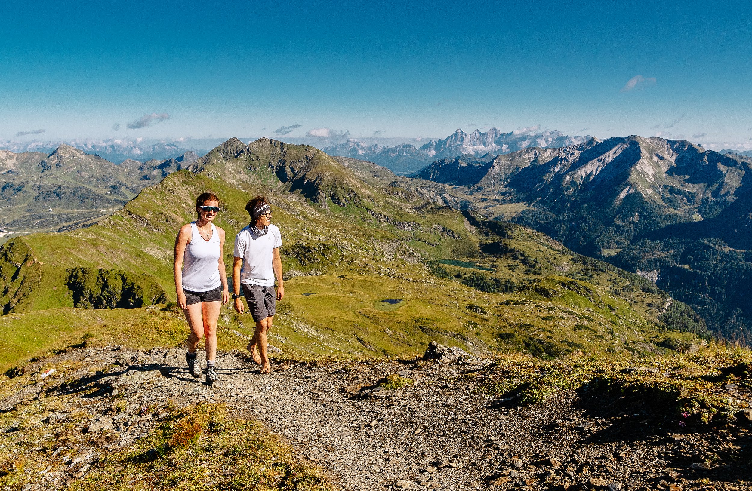 Zwei Wanderer auf einem Bergpfad in einer grünen, bergigen Landschaft mit hohen Gipfeln im Hintergrund bei sonnigem Wetter.
