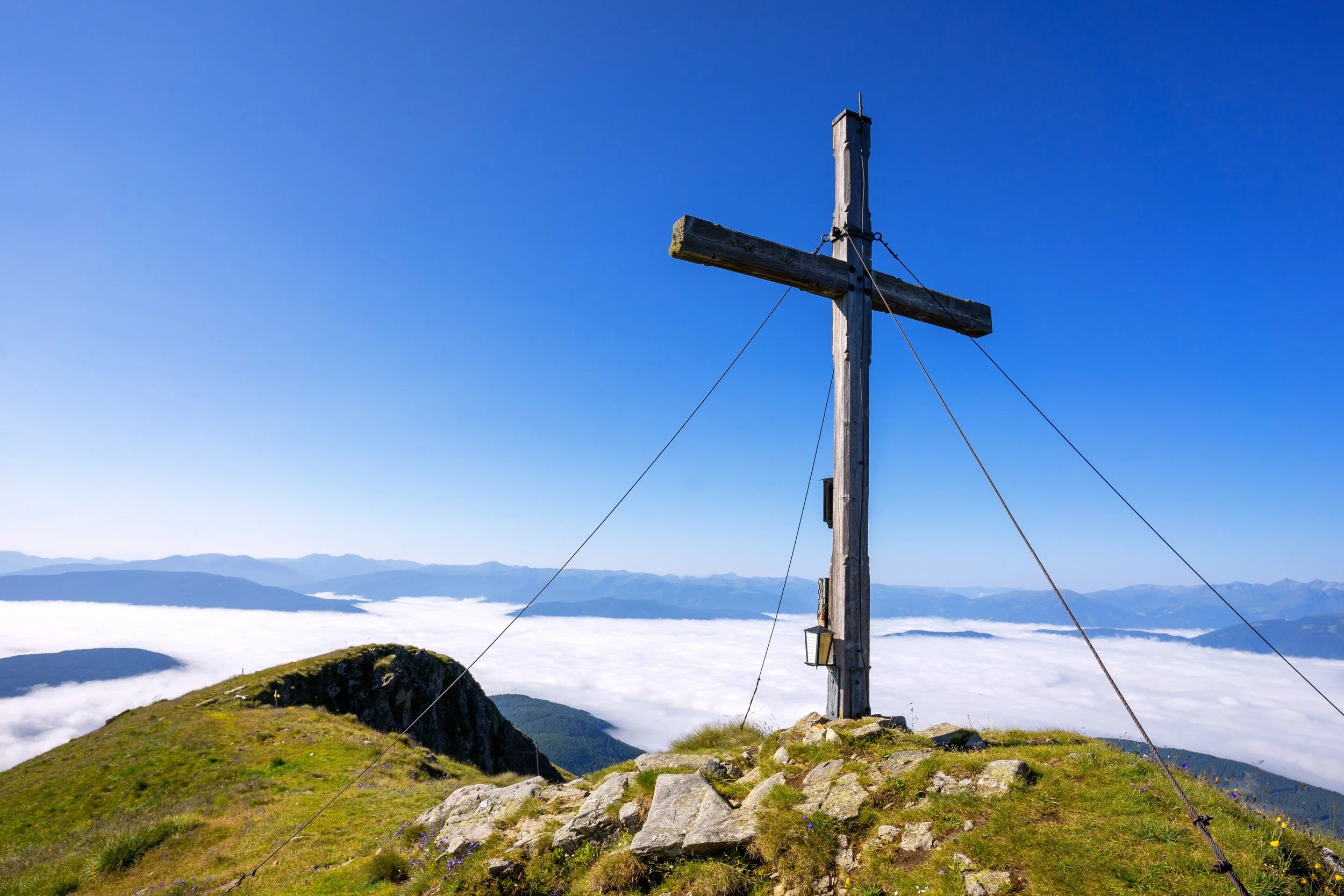 Ein Holz-Halbhorizont, das im Geiste eines Kreuzes gestaltet ist, steht auf einer mit Gras und Steinen bedeckten Bergspitze, mit Blick auf Wolken und Berge im Hintergrund unter klarem blauen Himmel.