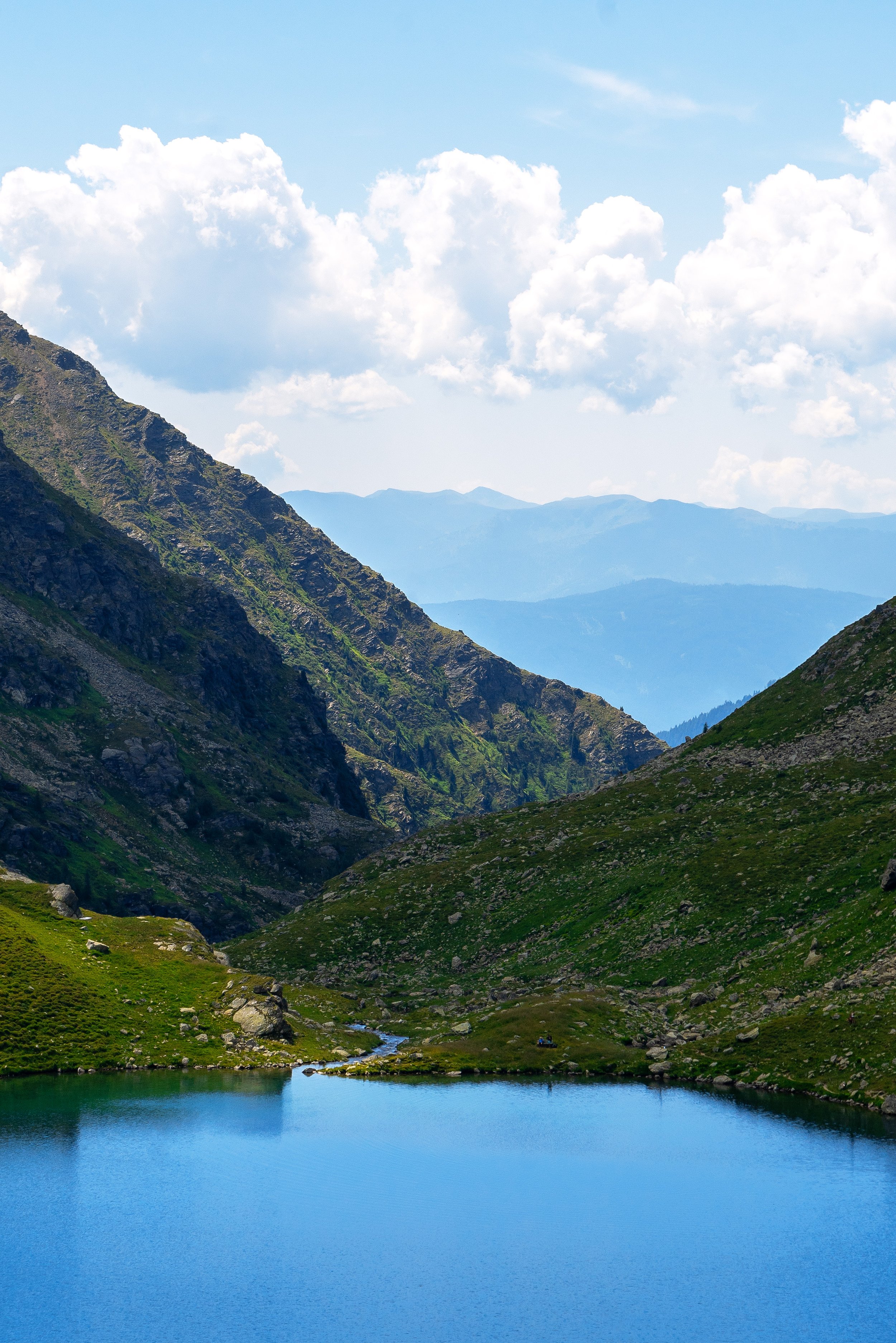 Bergsee in einer bergigen Landschaft mit grünen Hängen und Wolken am Himmel.
