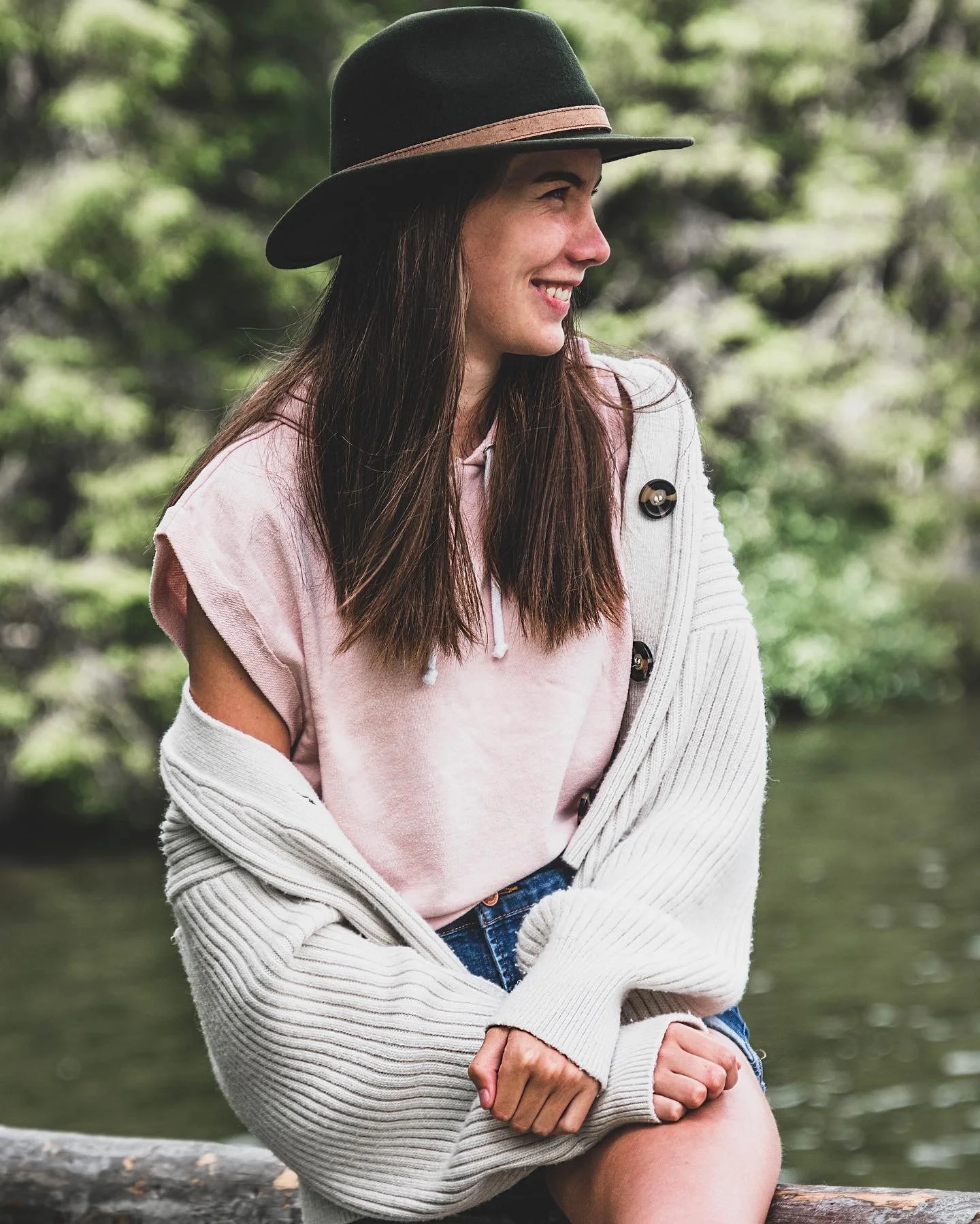 Junge Frau sitzt im Freien neben einem Fluss, trägt einen Hut, pinkes T-Shirt, graue Strickjacke, und lächelt.