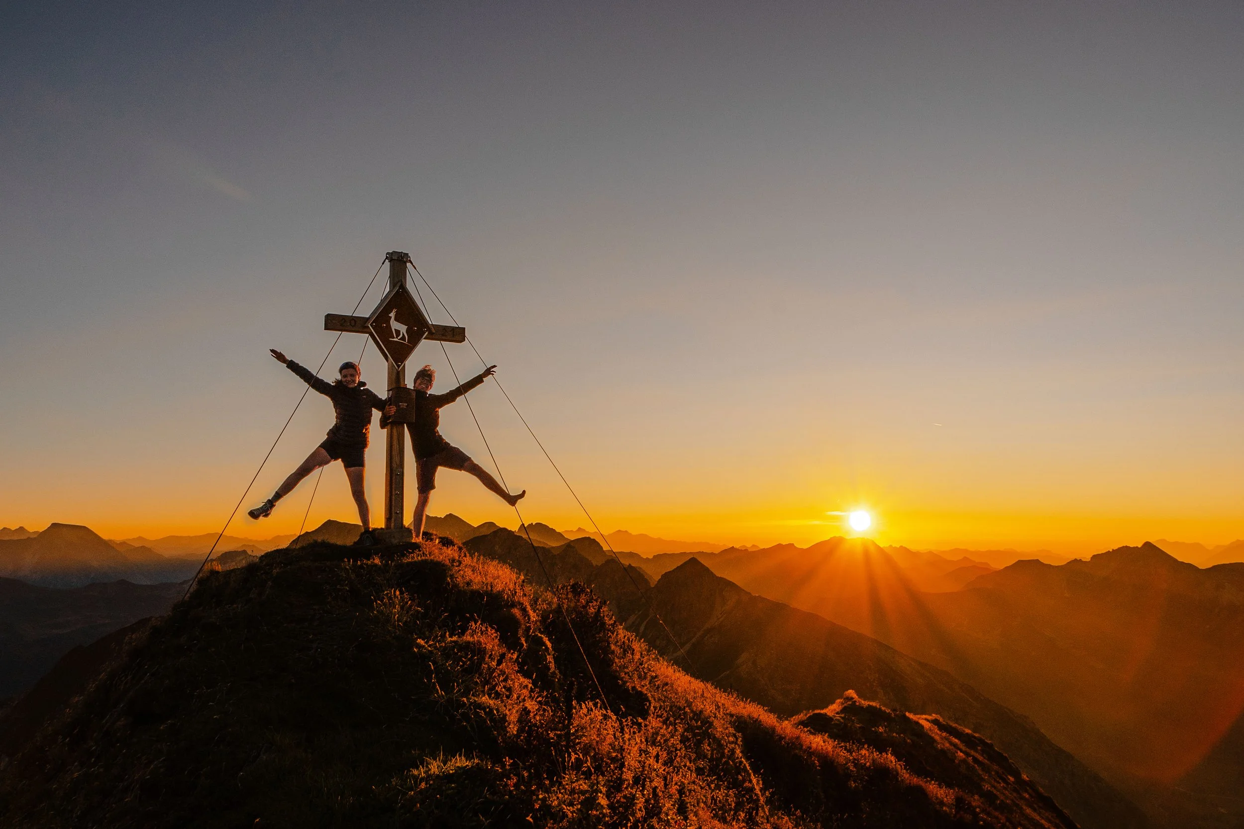 Zwei Personen auf einem Berggipfel bei Sonnenuntergang, die am Gipfelkreuz posieren, mit einer weiten Bergkulisse im Hintergrund.