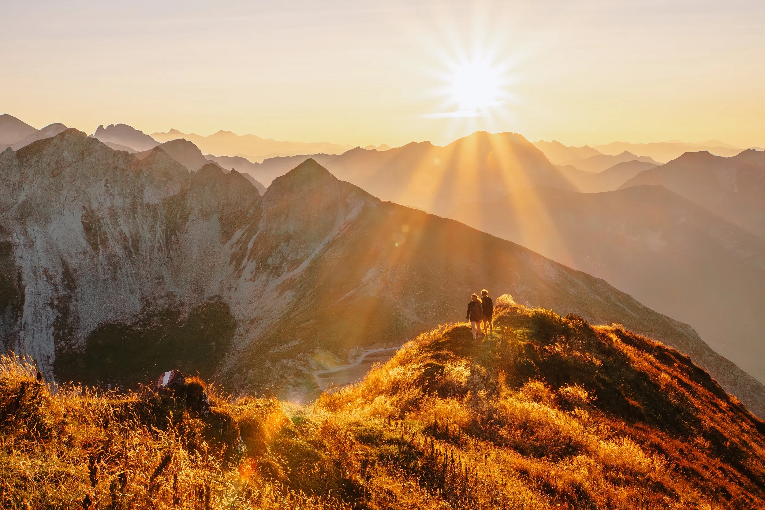 Zwei Wanderer auf einem sonnenverwöhnten Berggipfel bei Sonnenuntergang, umgeben von einer Bergkette und einem goldenen Himmel.