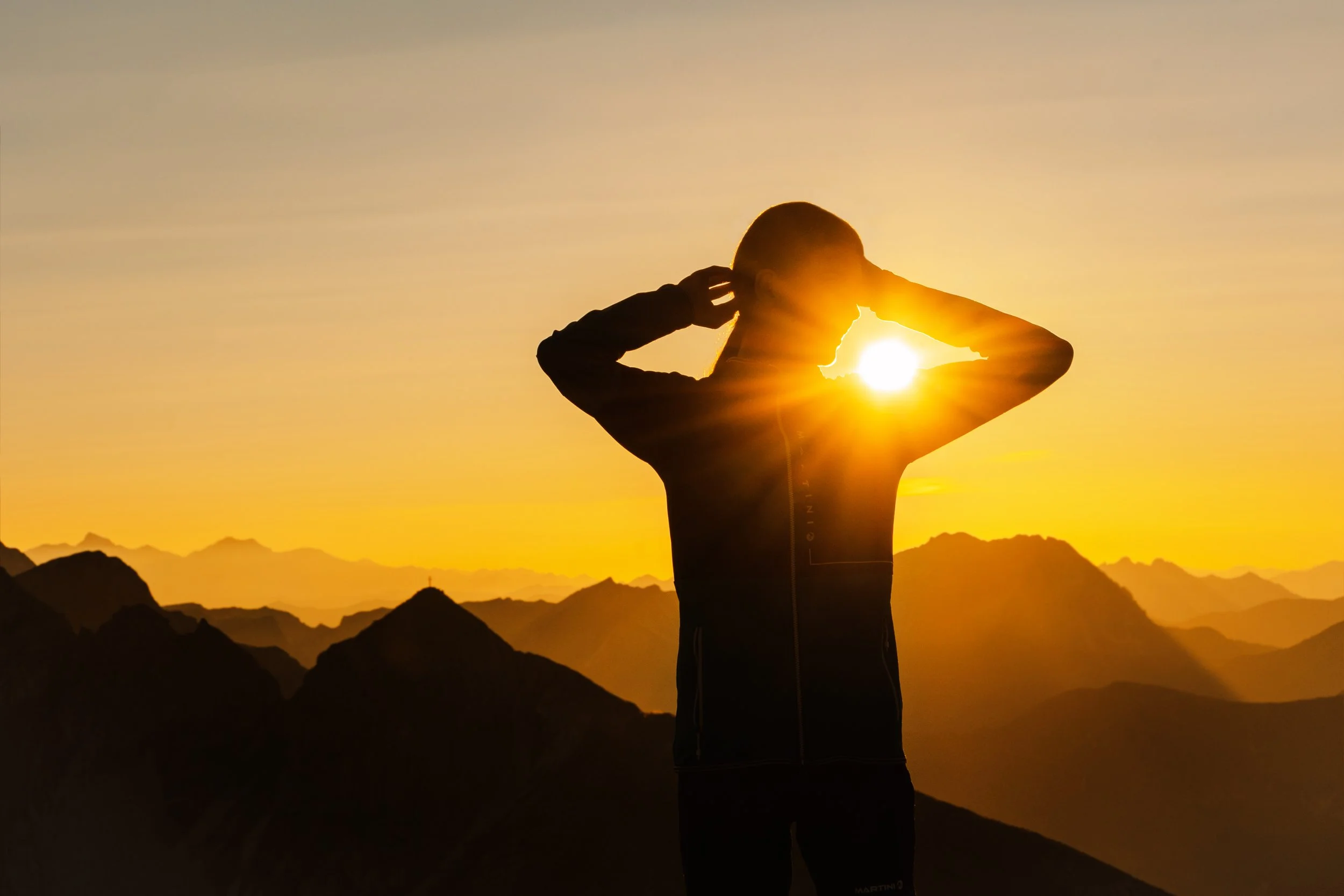 Eine silhouettierte Person, die den Sonnenaufgang oder Sonnenuntergang in einer bergigen Landschaft betrachtet, mit der Sonne, die durch ihre Arme scheint.