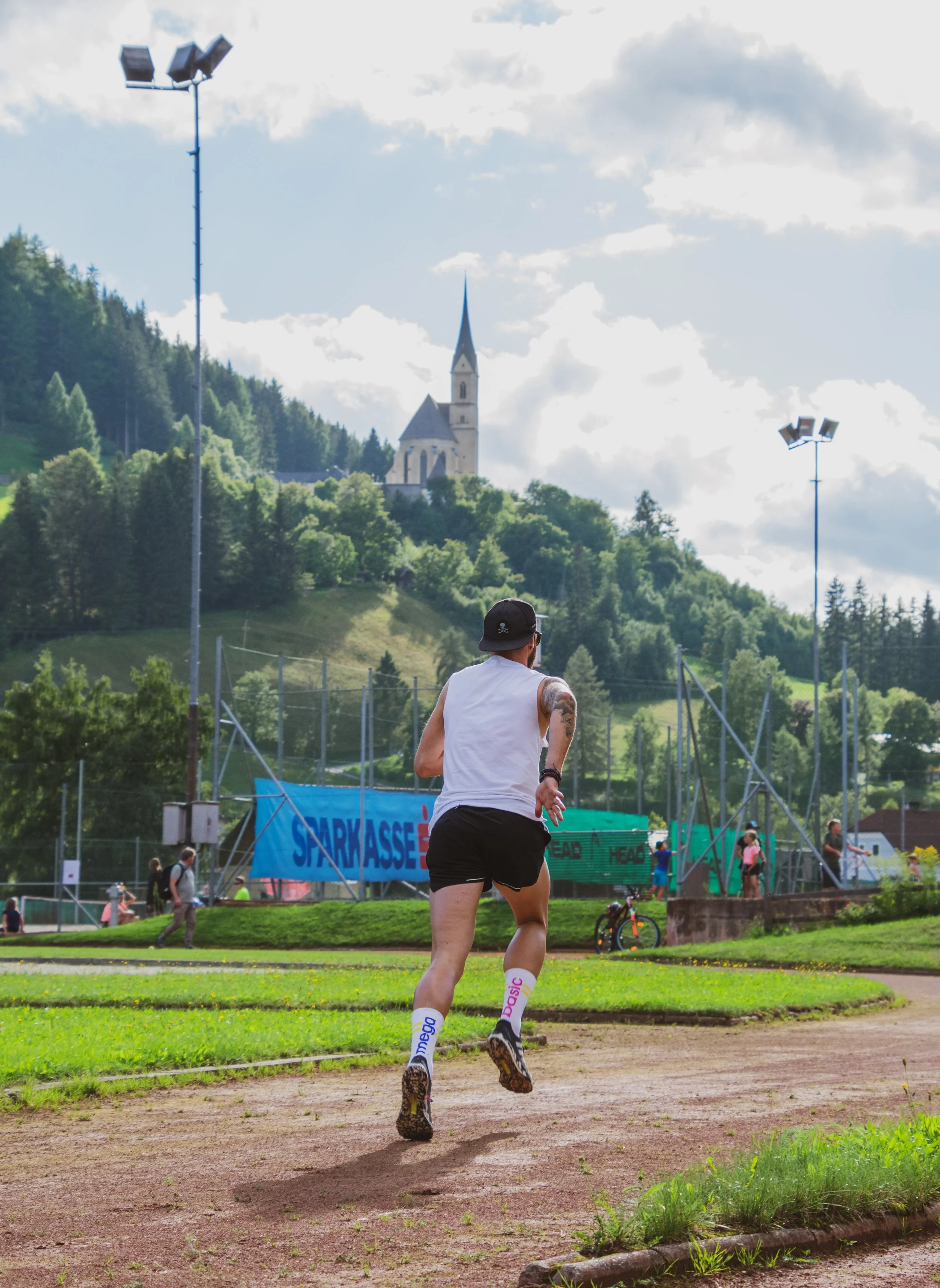 Ein männlicher Läufer in weißem Tanktop, schwarzer Shorts, Socken mit Markenname und einem schwarzen Cap läuft auf einer Laufbahn in einem Park im Freien, im Hintergrund befindet sich eine Tennishalle, Menschen und ein biblischer Turm auf einem Hügel