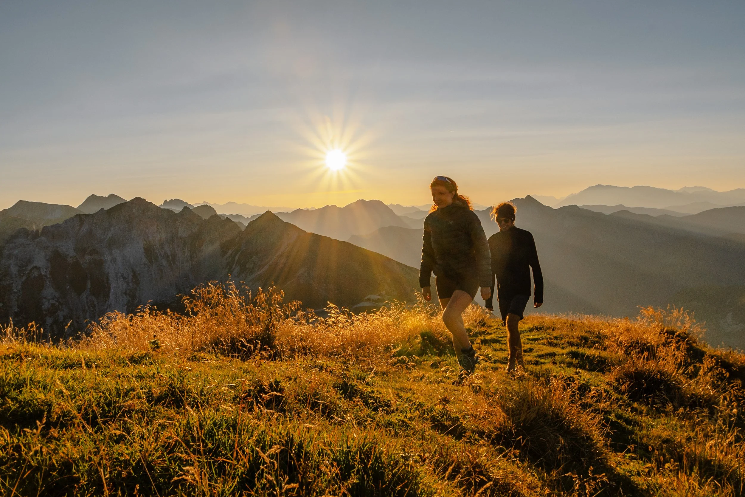 Zwei Wanderer in der Abendsonne auf einer Bergwiese mit Bergketten im Hintergrund.