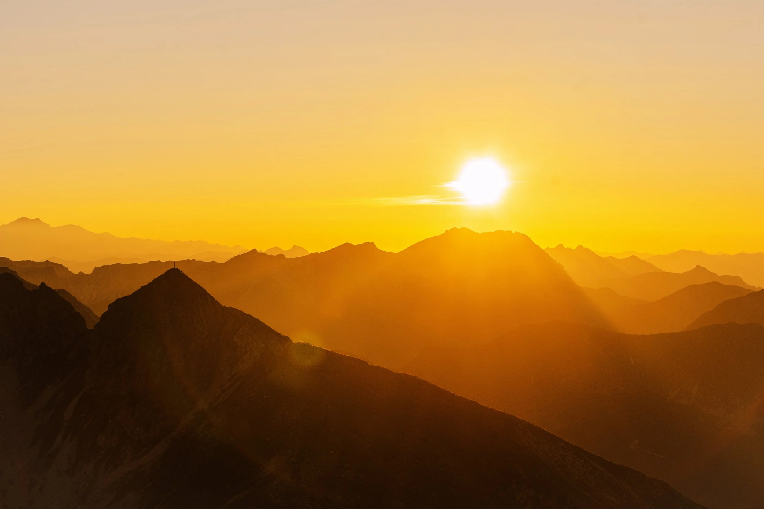 Sonnenuntergang über einem Gebirge mit mehreren schichten von Berggipfeln, die in warmen Orangetönen leuchten.