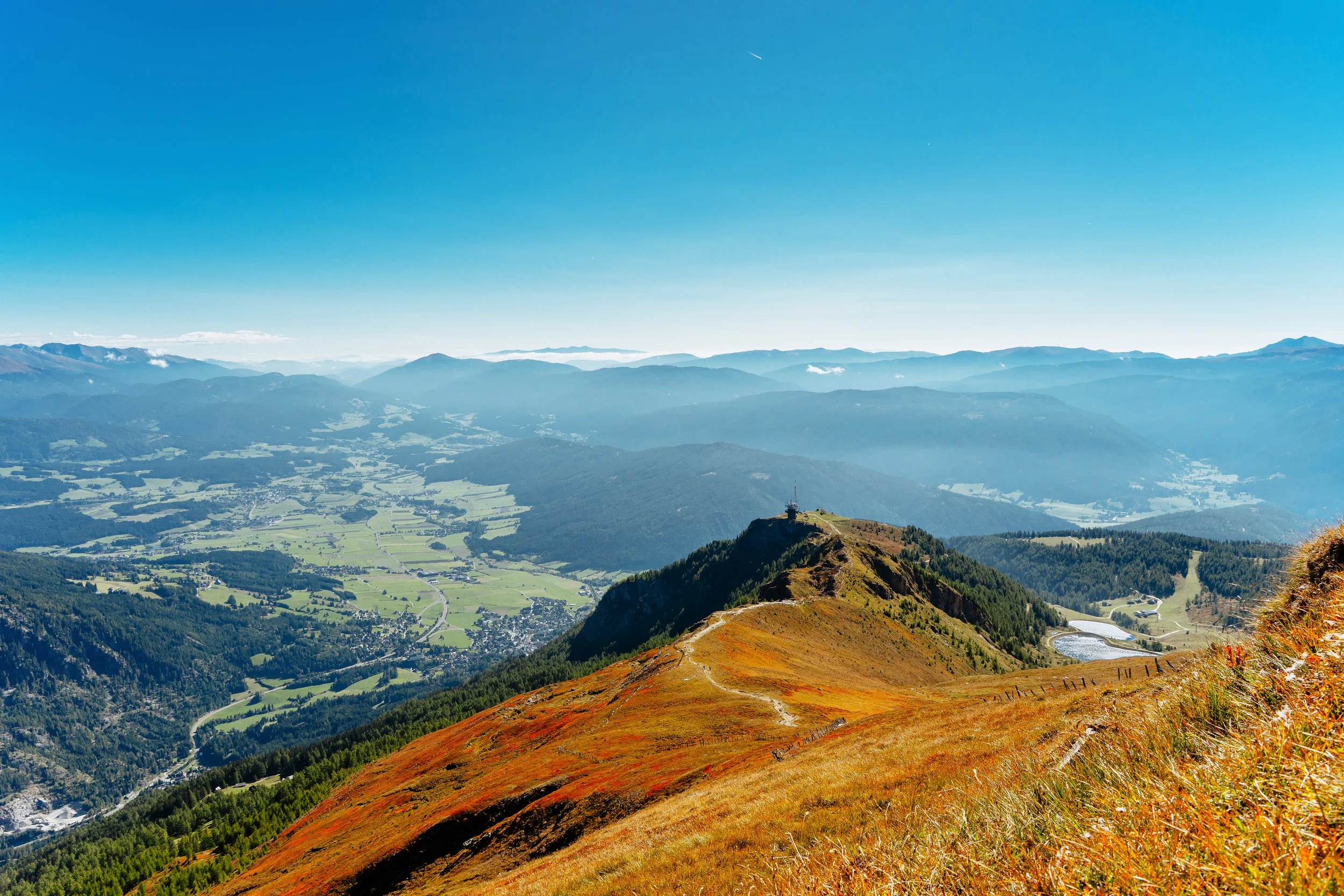 Blick auf eine bergige Landschaft mit einem Gipfel in der Mitte, umgeben von Wäldern, Wiesen und einer tief liegenden Dörfer in der Ferne unter blauem Himmel.
