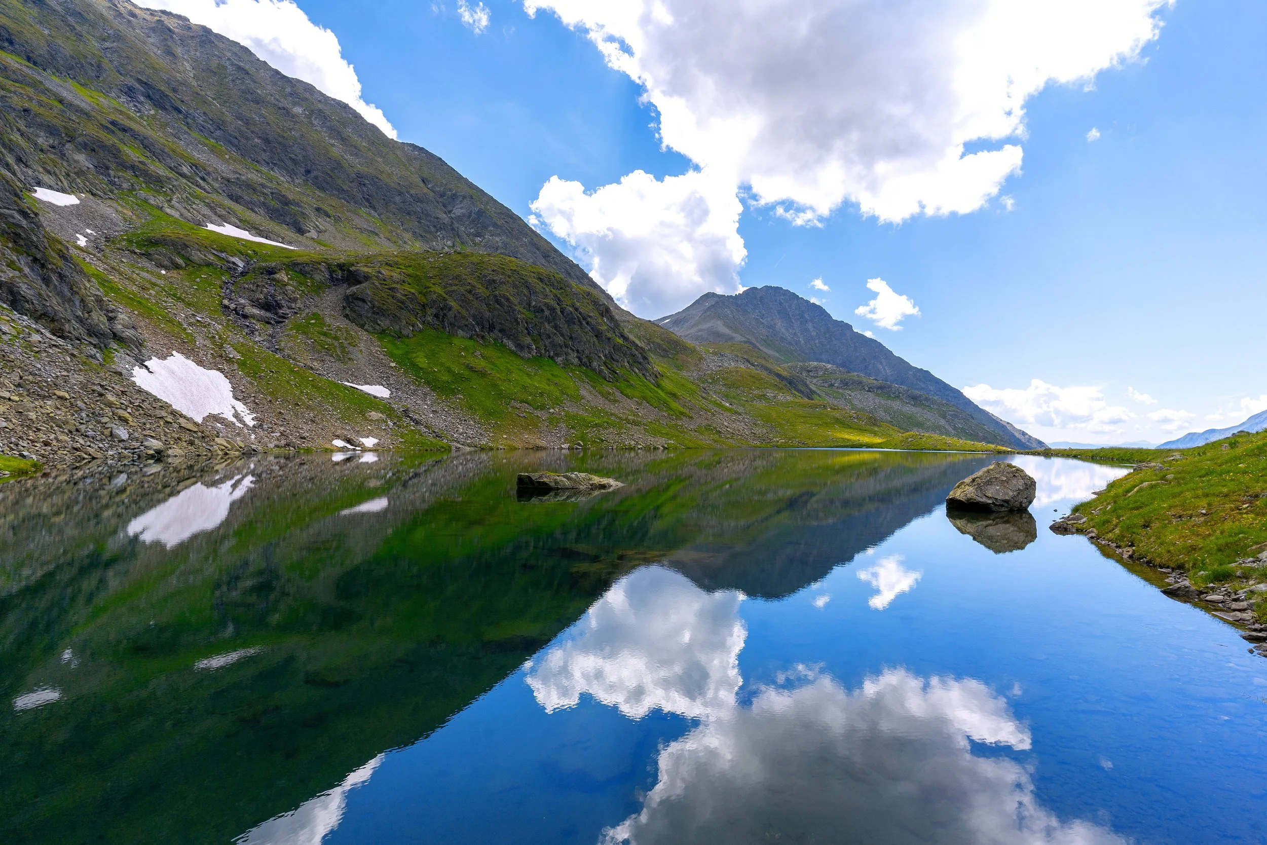 Bergsee in einer Berglandschaft mit Wasser, das die Wolken und Berge reflektiert, umgeben von grünen Wiesen und felsigen Hügeln, unter einem blauen Himmel mit einigen Wolken.