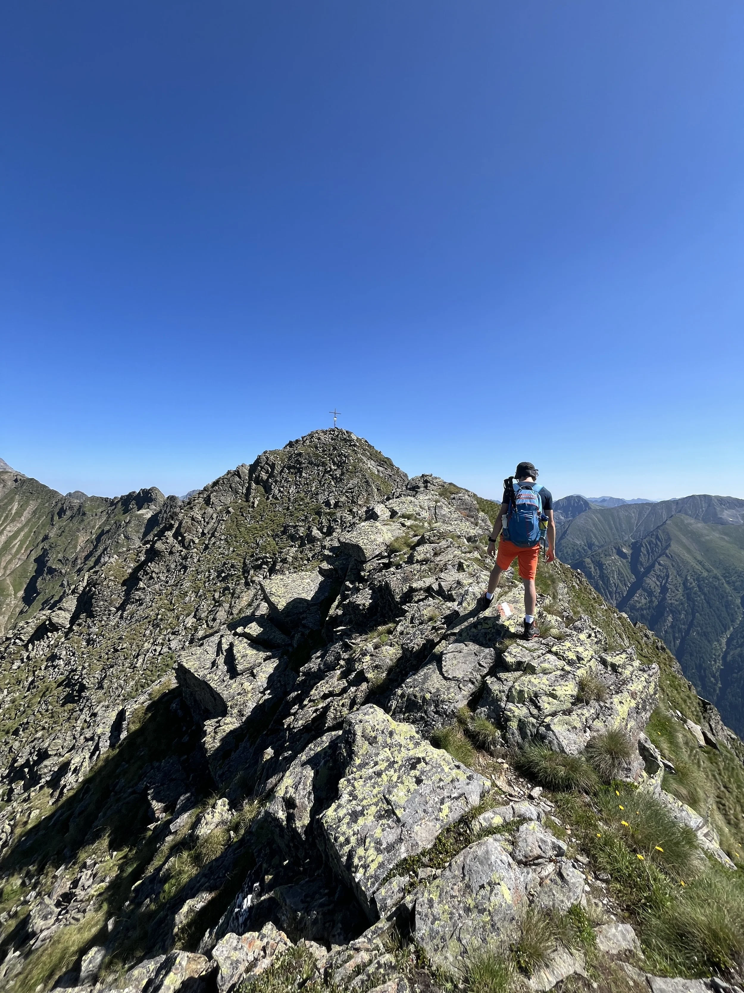 Ein Wanderer mit Rucksack auf einem schmalen, felsigen Gebirgskamm unter klarem blauen Himmel. Im Hintergrund ist eine Bergkuppe mit einem Gipfelkreuz zu sehen.