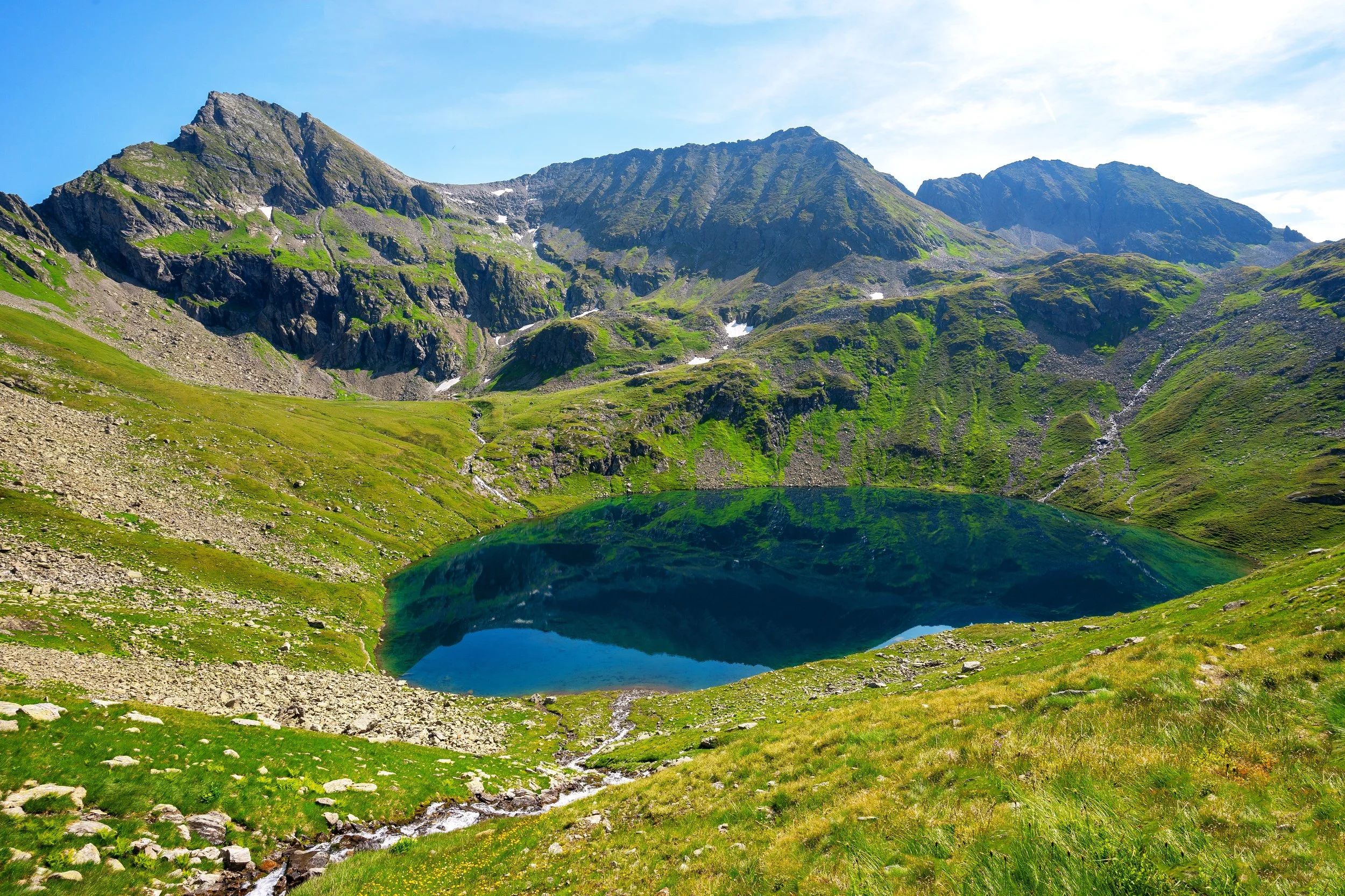 Bergsee inmitten grüner Berge und hoher Berge im Hintergrund bei sonnigem Himmel.