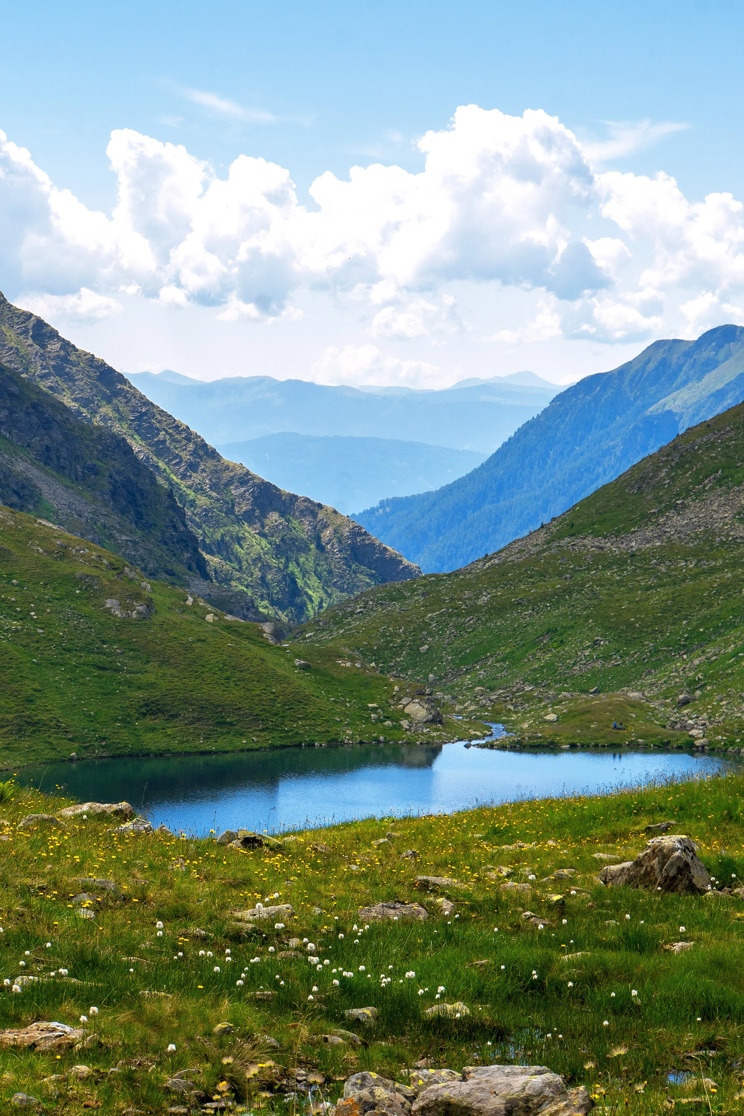 Bergsee in einer grünen, bergigen Landschaft mit Wolken am Himmel.