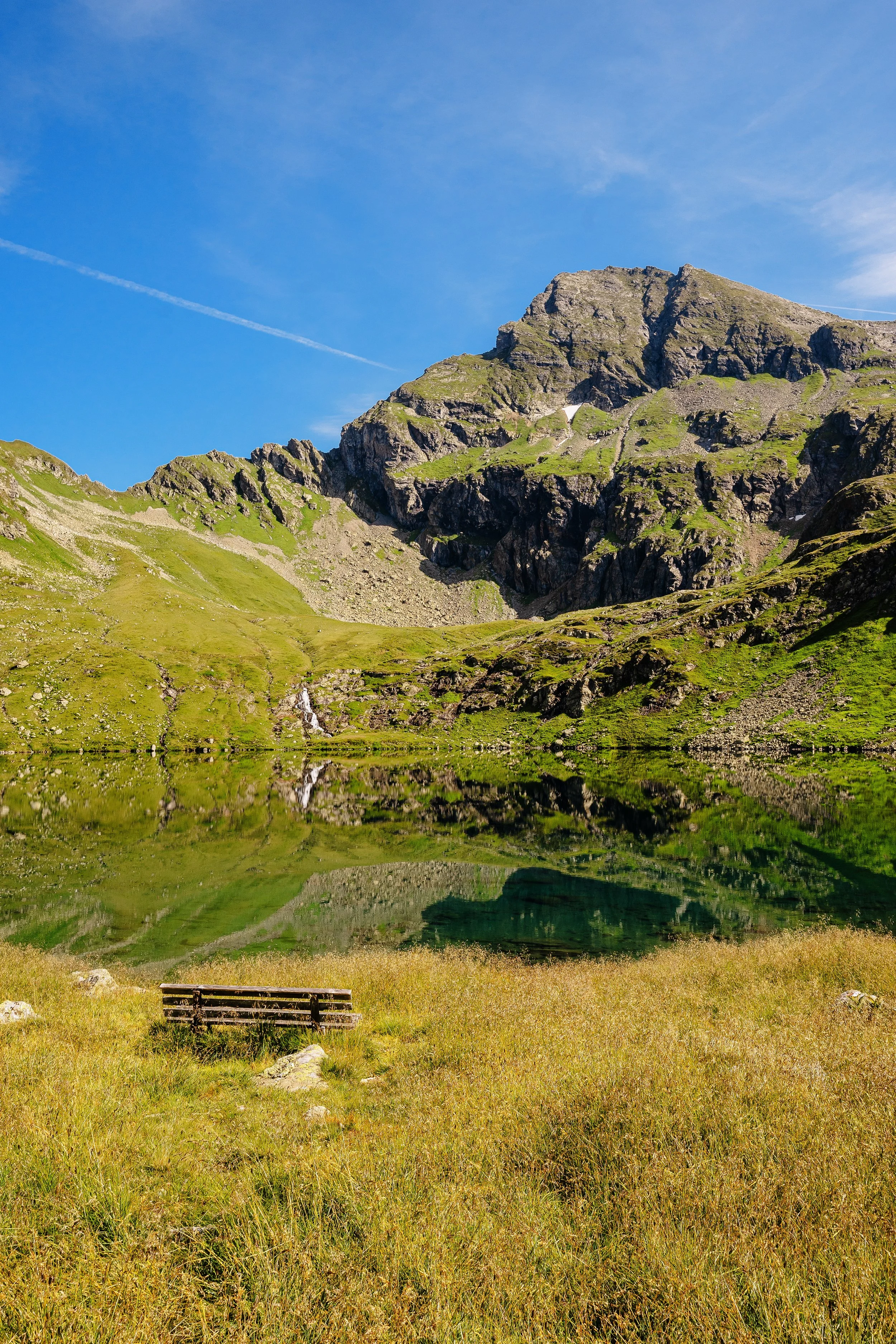Berge, ein See, eine grüne Wiese mit einer Bank und ein blauer Himmel.