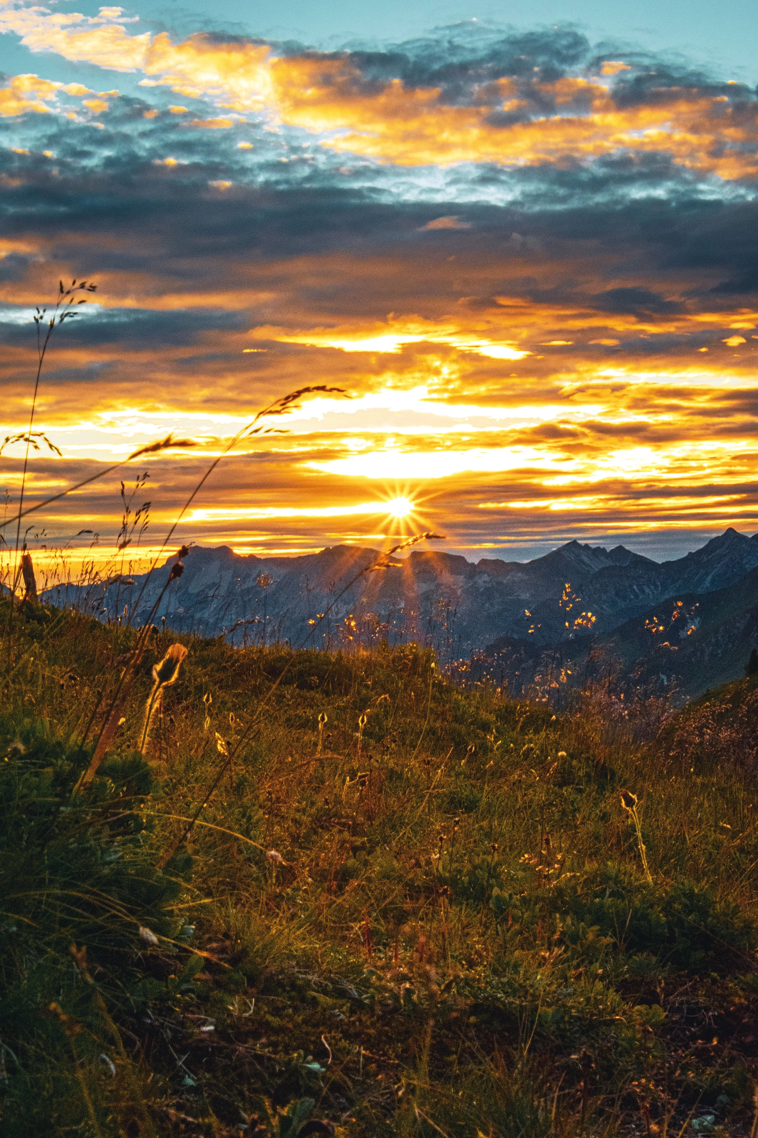 Ein malerischer Sonnenuntergang über Bergen, die im Hintergrund sichtbar sind, während das Licht die Wolken und die Wiese im Vordergrund beleuchtet.