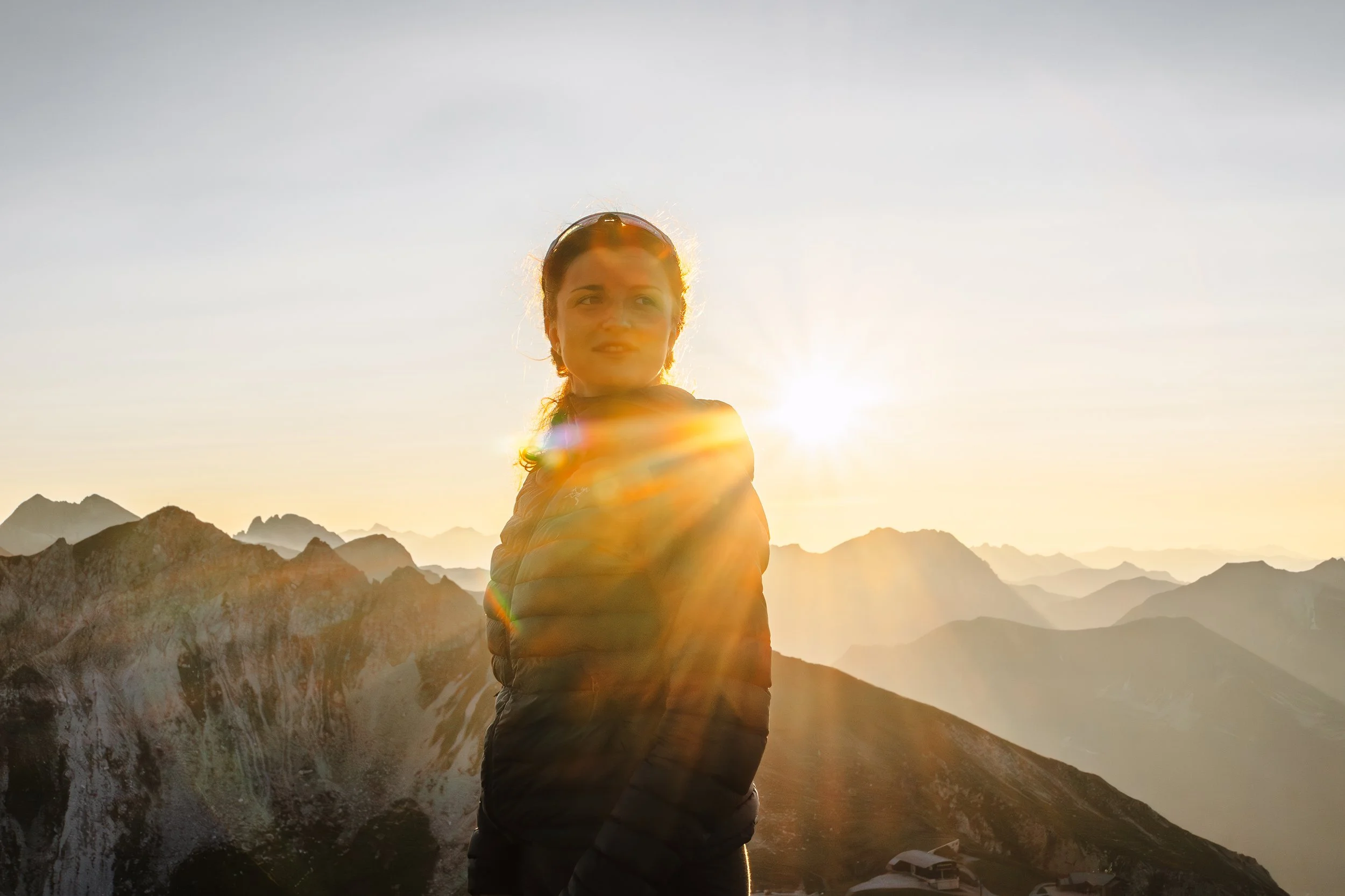 Junge Frau in dunkler Jacke vor Bergen bei Sonnenuntergang, Sonnenstrahlen hinter ihr.