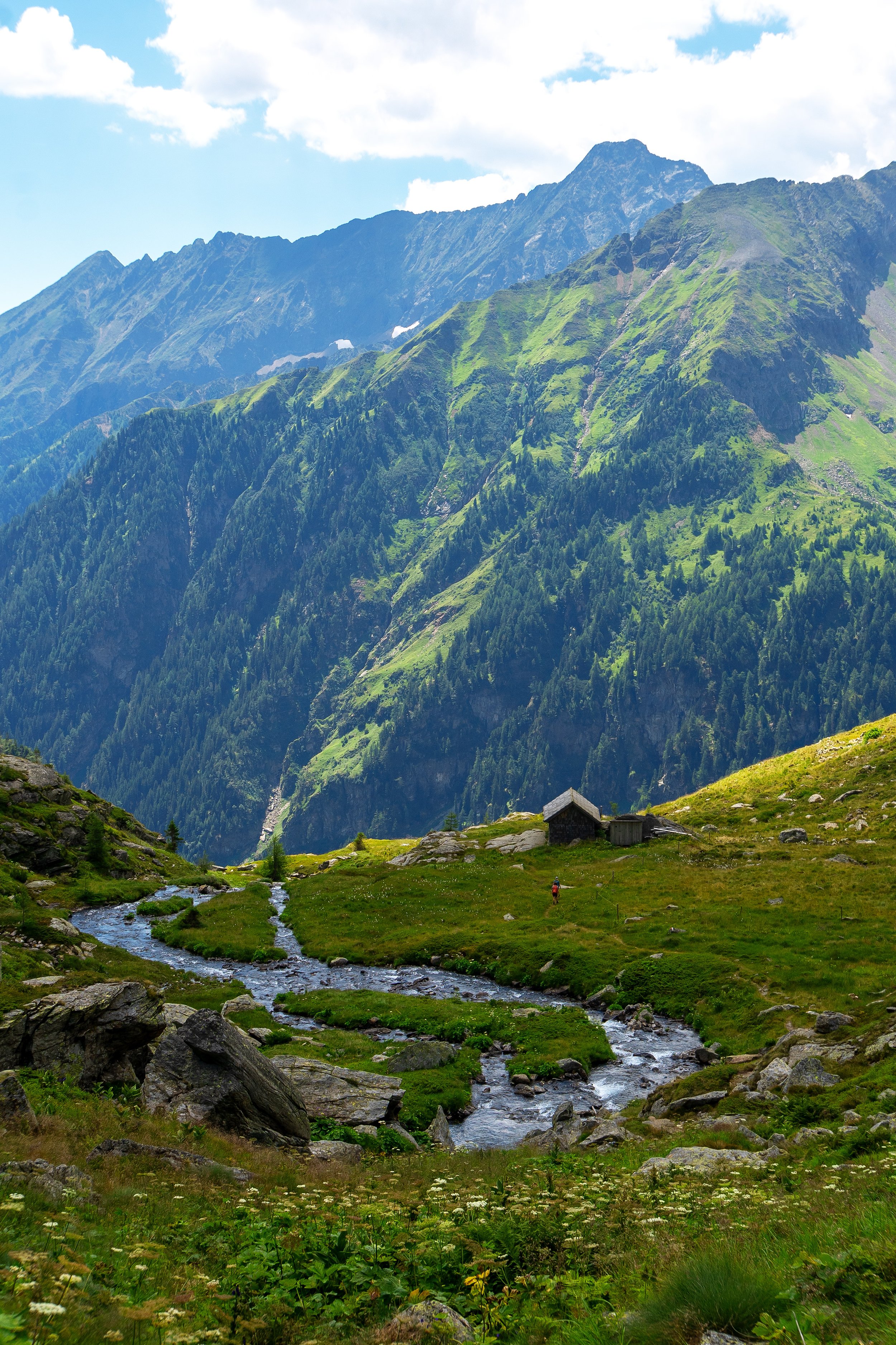 Berglandschaft mit grünem Tal, fließendem Bach, Holzhaus, Wanderer und hohen Bergen im Hintergrund unter einem bewölkten Himmel.