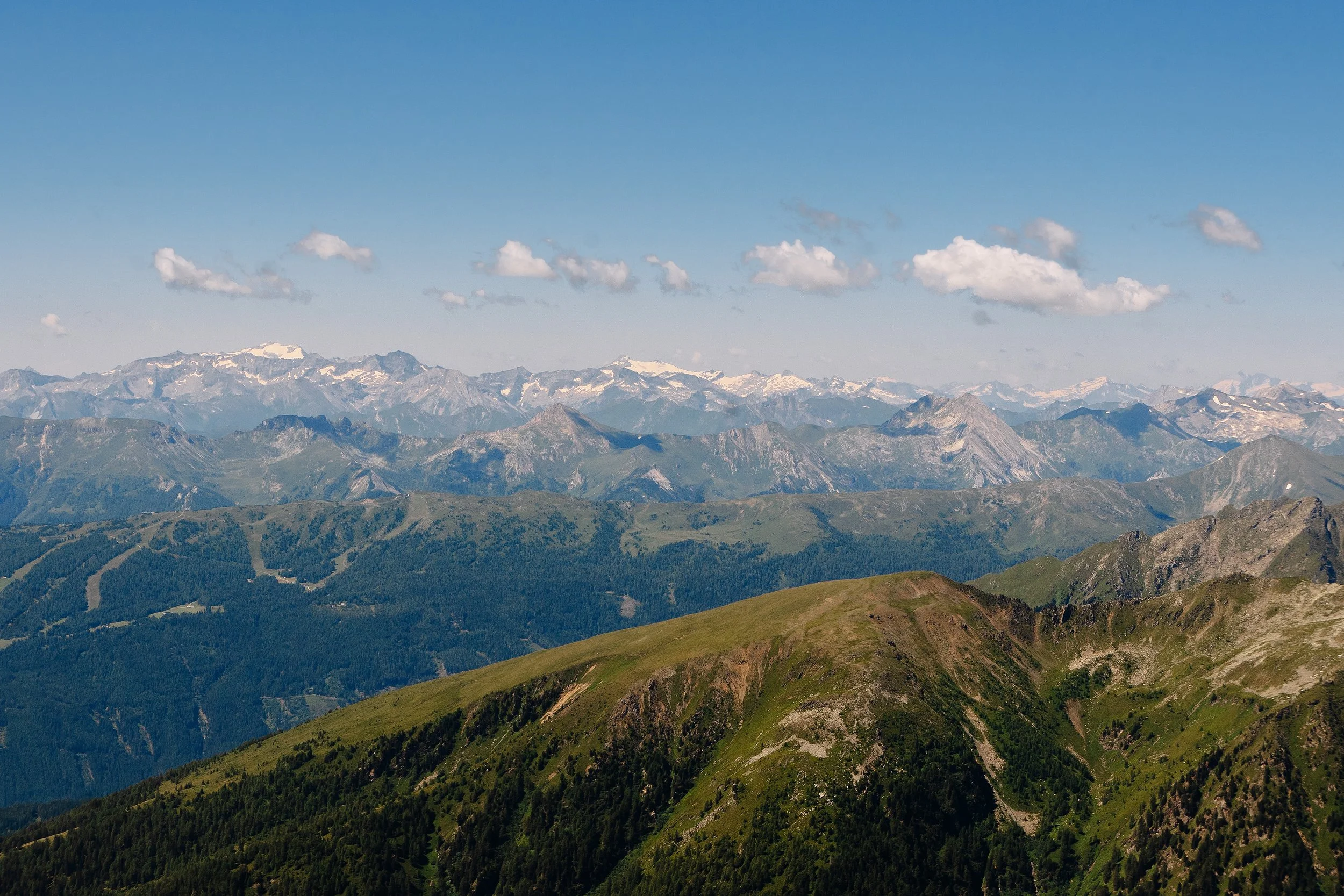 Bergpanorama mit grünen Hügeln im Vordergrund, höheren Bergen im Hintergrund und einen blauen Himmel mit einigen Wolken.