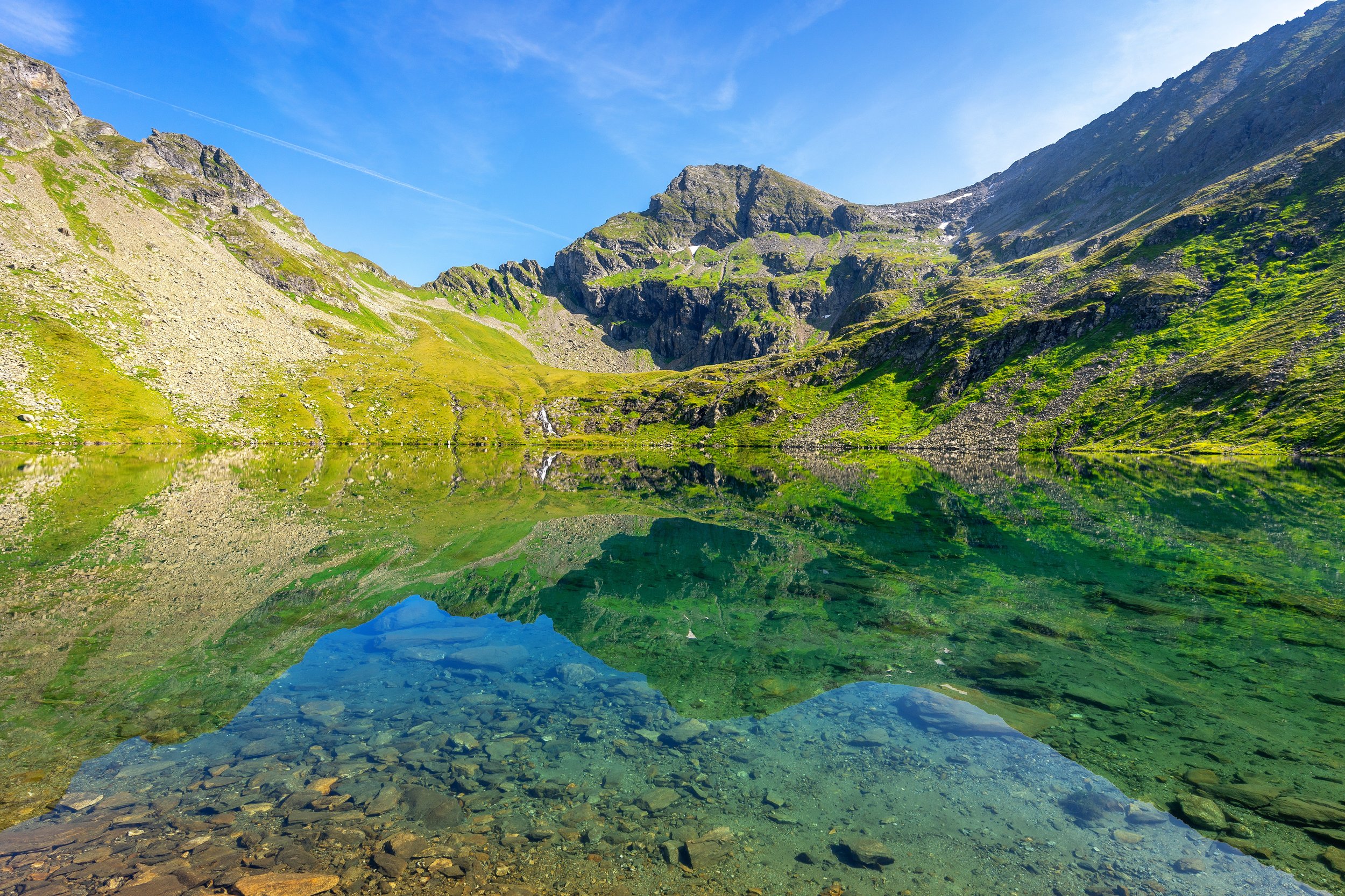 Bergsee inmitten grüner Berge unter blauem Himmel, mit Reflexion der Berge im Wasser.