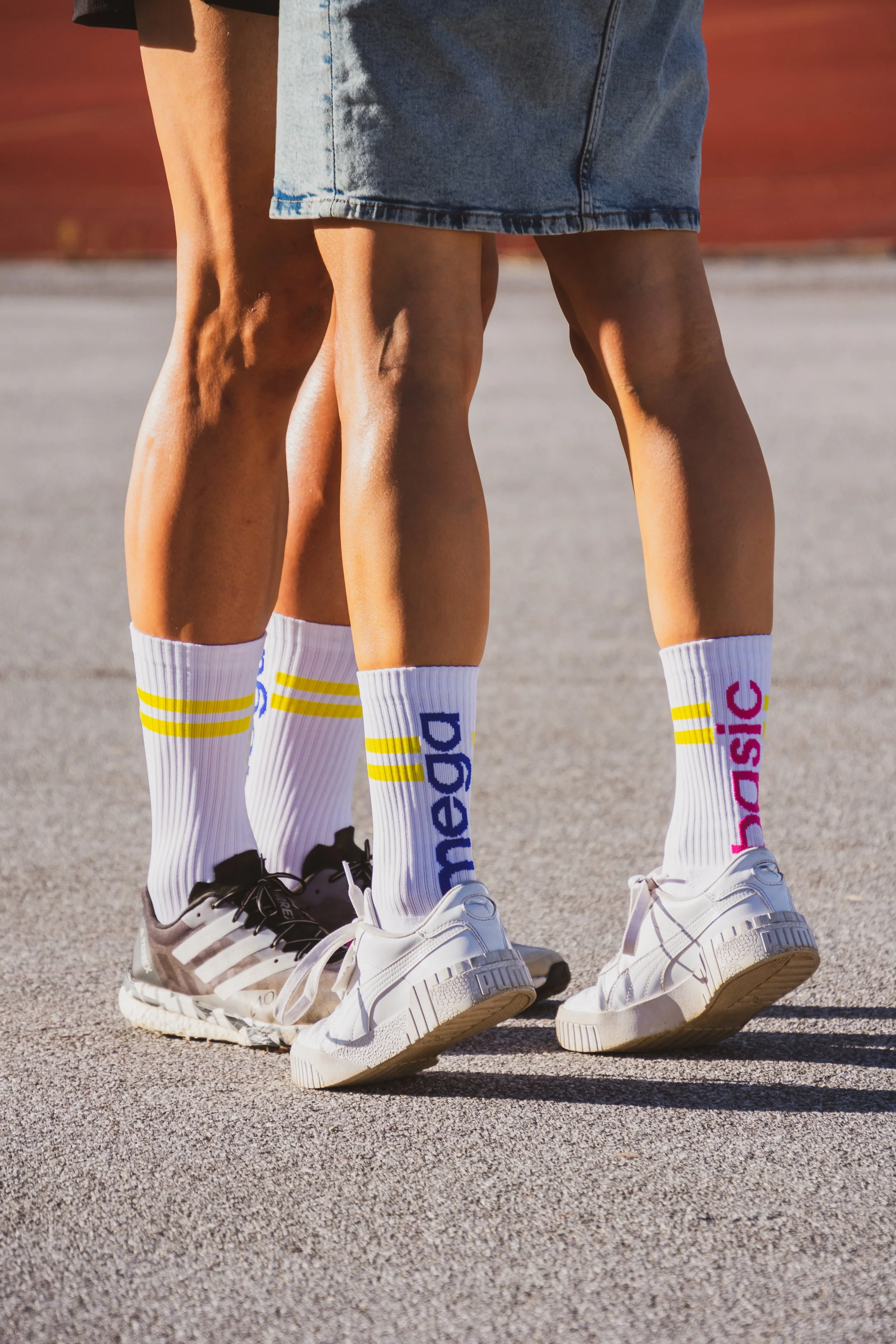 Nahaufnahme von zwei Menschen, die auf einer Asphaltstraße stehen, mit sichtbaren Beinen, weißen Adidas-Sneakers und bunten Socken mit der Aufschrift 'adidas'.