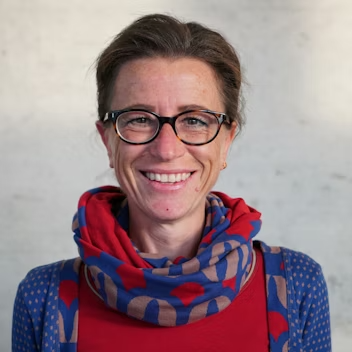 A woman with short brown hair, glasses, and a bright smile, wearing a red and blue patterned scarf and a blue polka dot shirt, standing against a plain light background.
