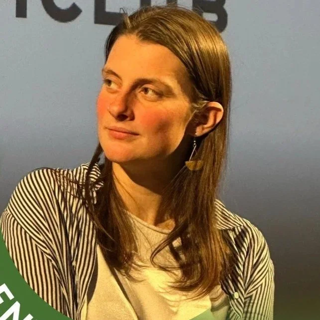 Adele Zambaldi, a woman with long brown hair looking to her left, wearing a striped cardigan and a light shirt, standing in front of a blue background.