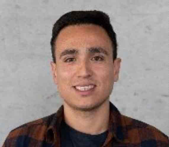 Diego Gonzalez, a smiling man with short dark hair, wearing a plaid shirt over a dark t-shirt, standing against a plain light-colored wall.