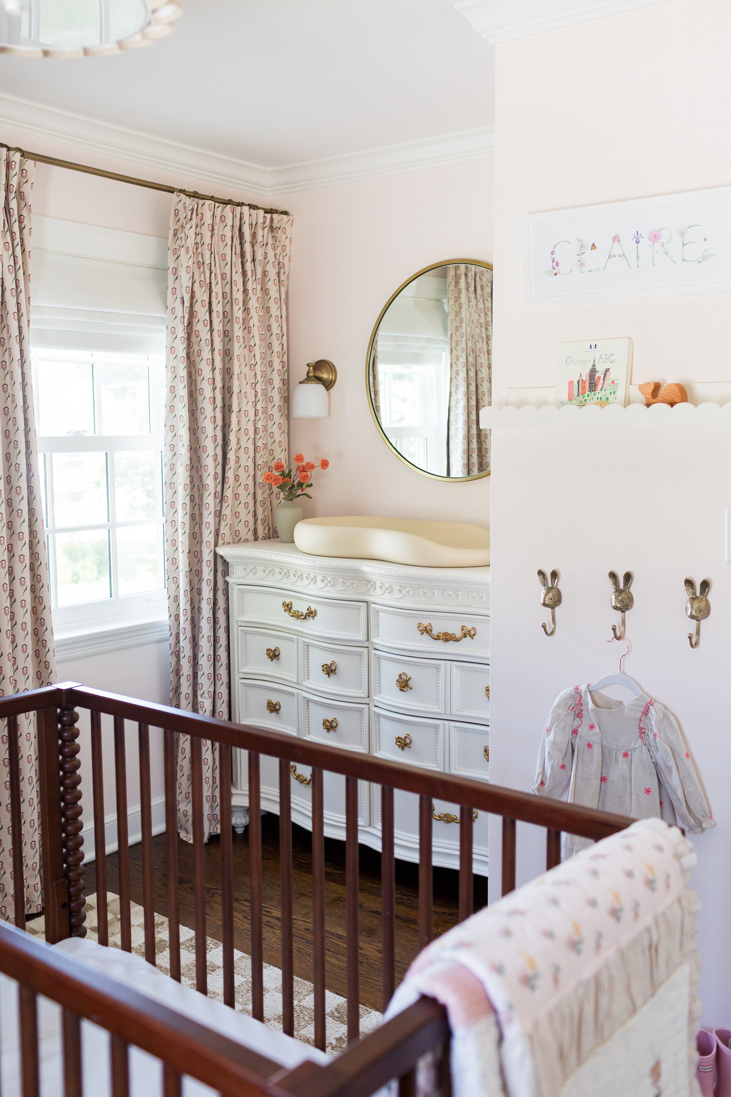 A nursery with a wooden crib, a white dresser with gold handles, a mirror, curtains with a pink and white pattern, a wall lantern, a vase with pink flowers, a framed art piece with the name 'Claire' decorated with flowers, and a small hanging girl’s 