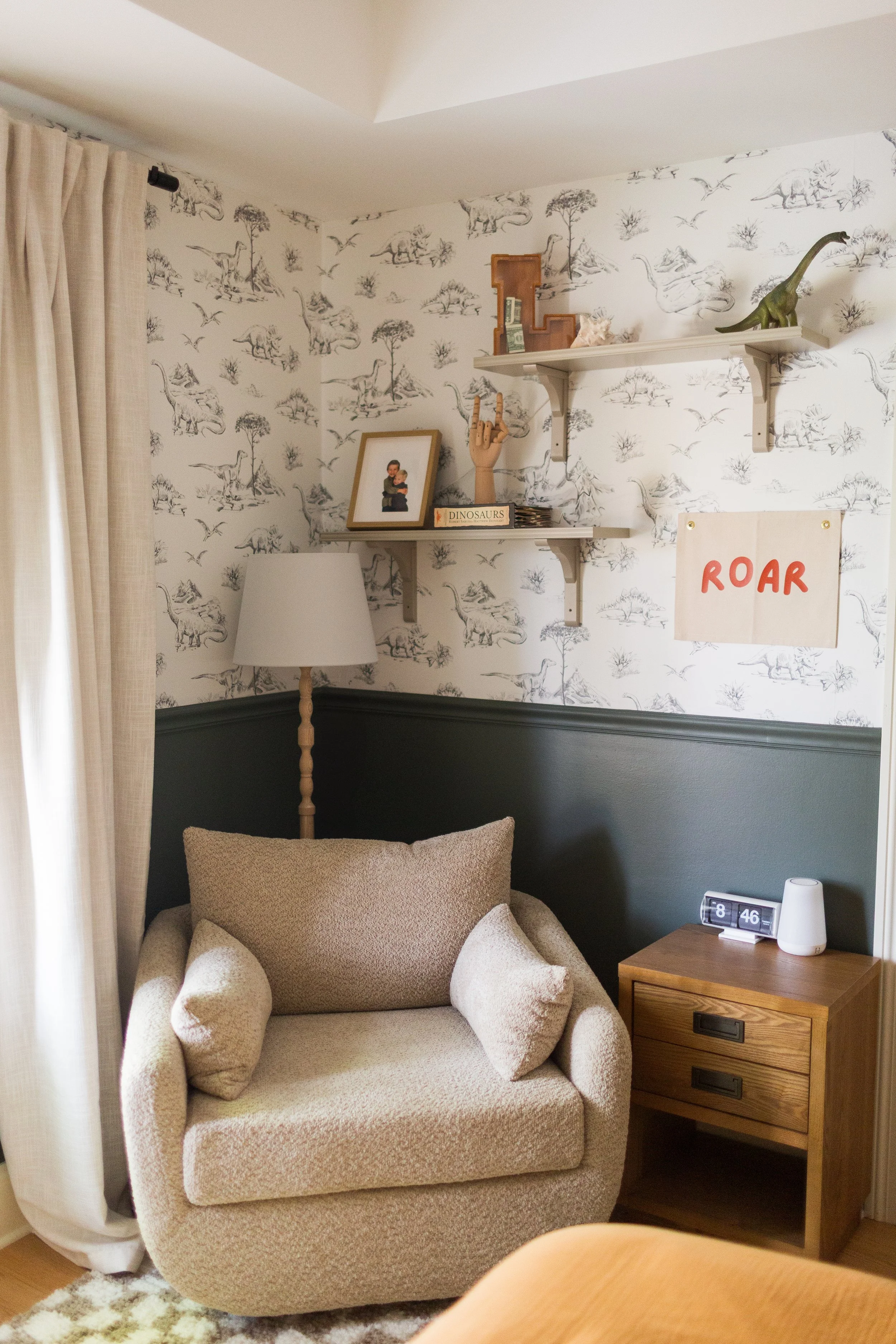 Living room corner with beige armchair, side table, wall shelf with dinosaur decorations, framed photo, and a lamp, with patterned wallpaper and beige curtains.