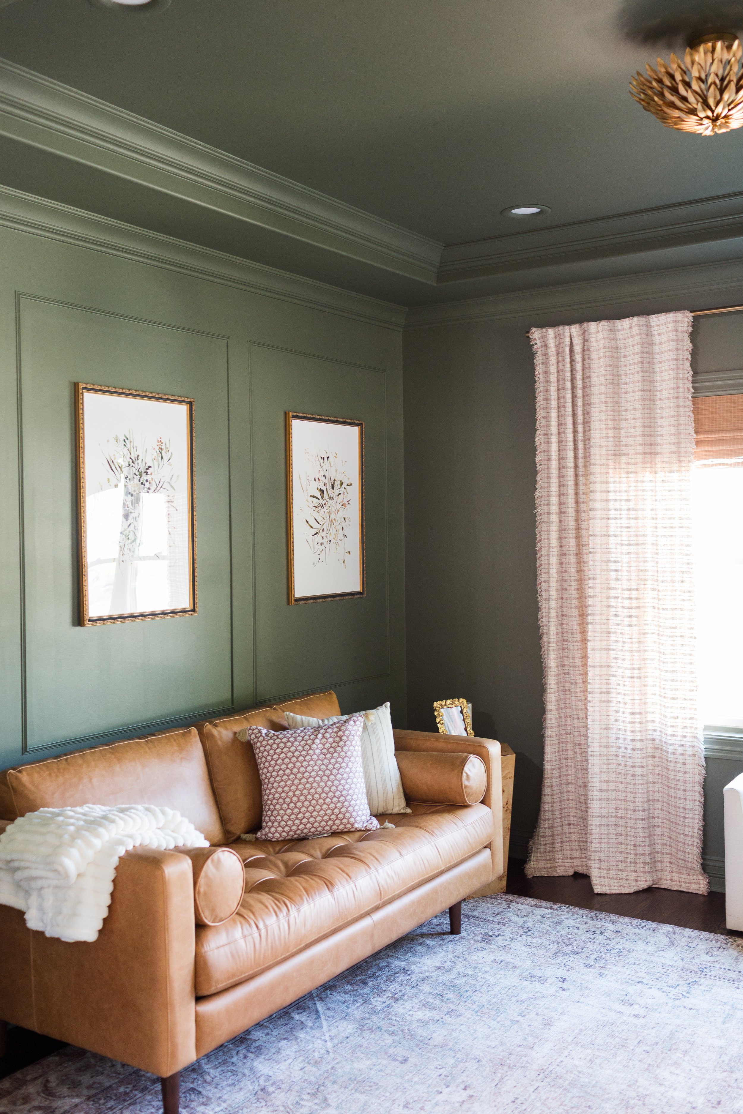 A living room with a tan leather sofa decorated with patterned pillows, a white blanket, and a rug, against a green wall with framed botanical artwork, and a window with pink checked curtains.