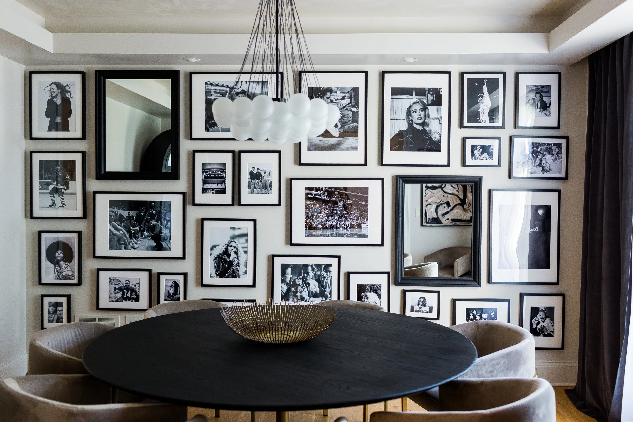 A dining room with a black round table surrounded by beige upholstered chairs, a large chandelier with white globes hanging from the ceiling, and a wall decorated with black and white photographs of various people.