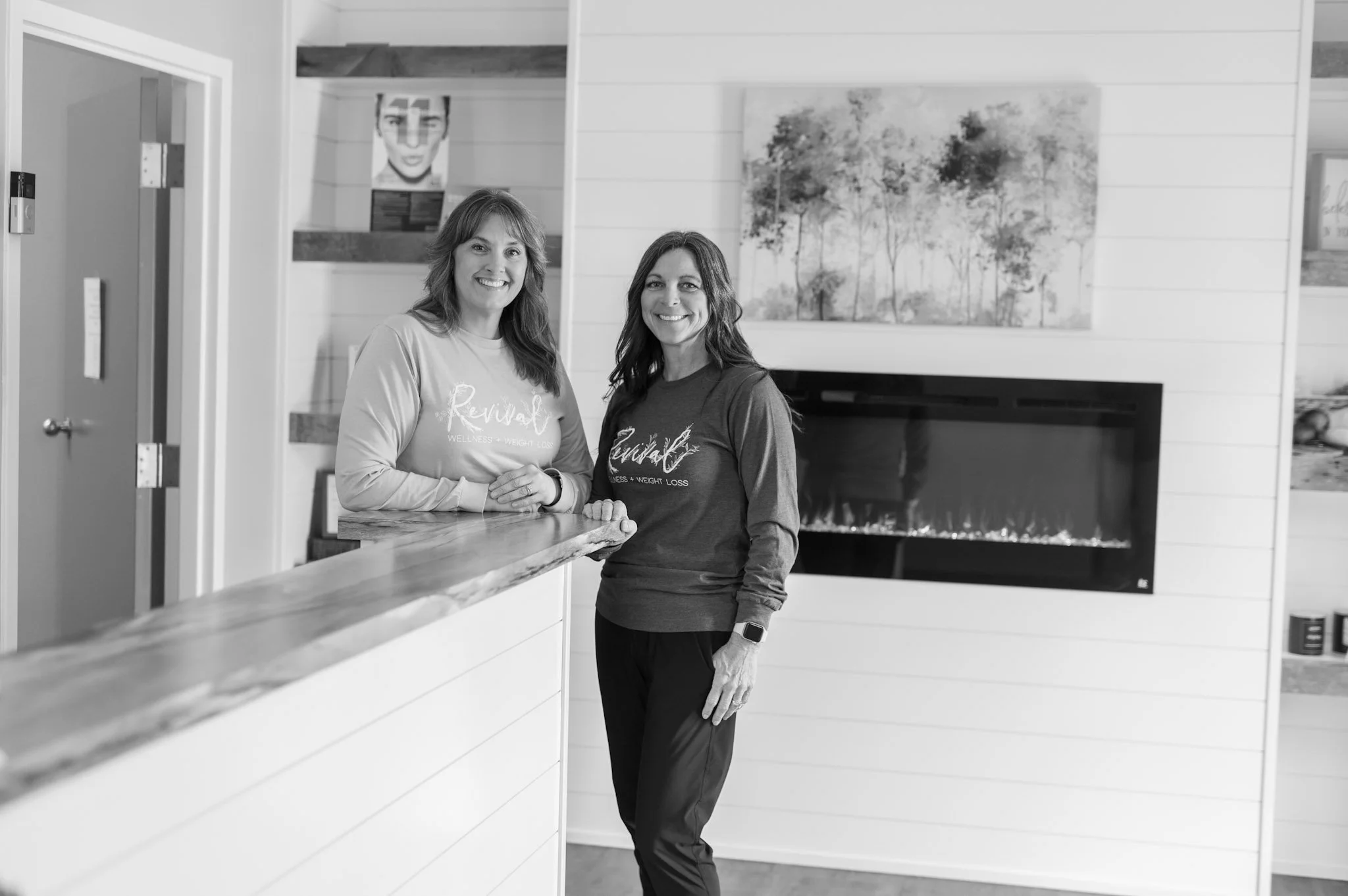 Two women smiling, standing next to each other inside a modern, decorated room with floating shelves, artwork, and plants.