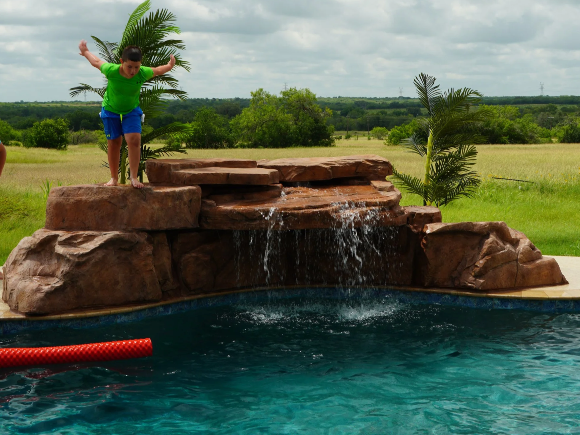 Kid jumping off an artificial pool waterfall into some blue water