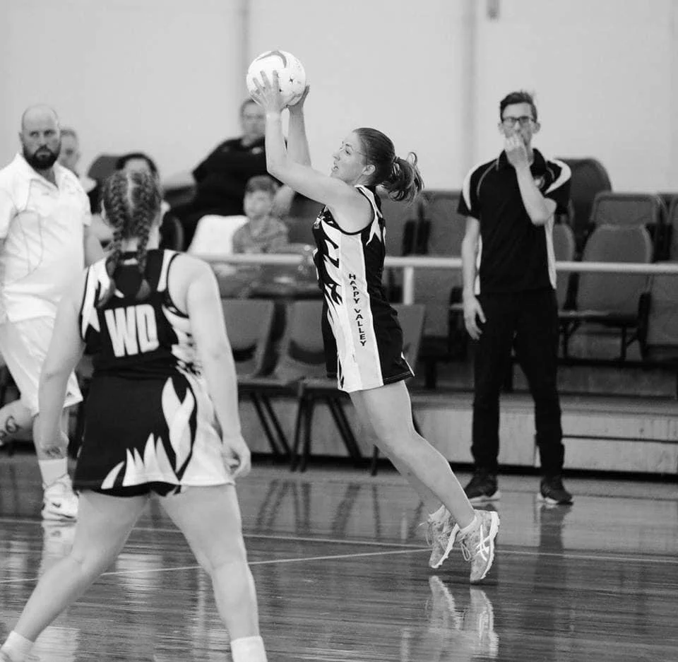 Black and white photo of a netball game featuring players in action, including a jumping player catching the ball. A spectator and coach watch from the sidelines. Players wear uniforms with names printed on the sides.