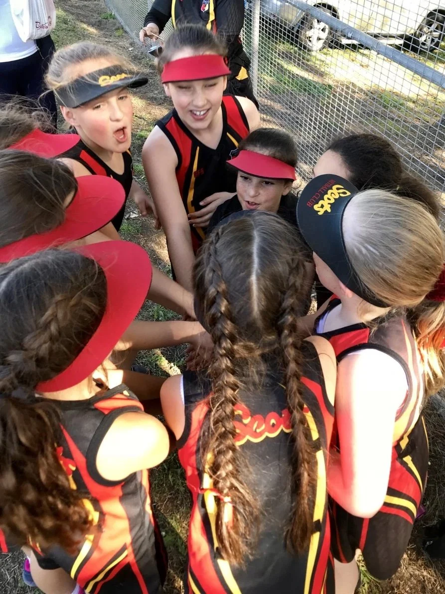 Young girls in sports uniforms and visors in a huddle outdoors, showing team spirit.
