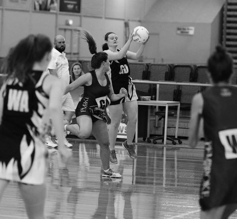 Netball game with players in motion inside a gymnasium, featuring a player catching the ball.