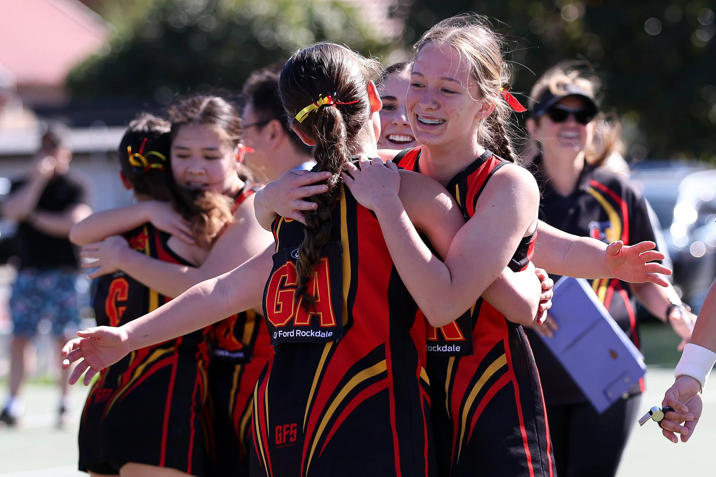 Netball players in red and black uniforms celebrating, hugging on an outdoor court, smiling and showing excitement.