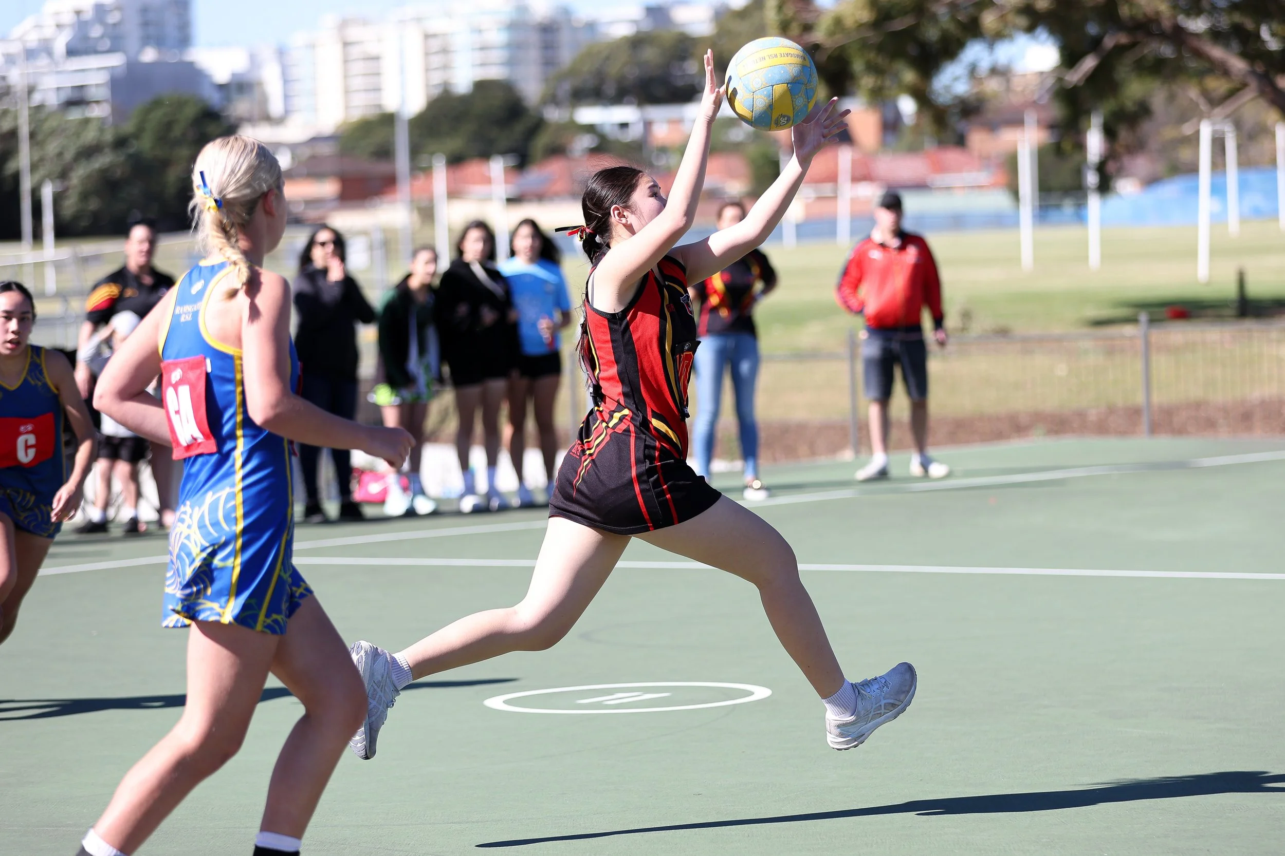 Netball game with players in blue and black uniforms on an outdoor court, one jumping to catch the ball, spectators in background.