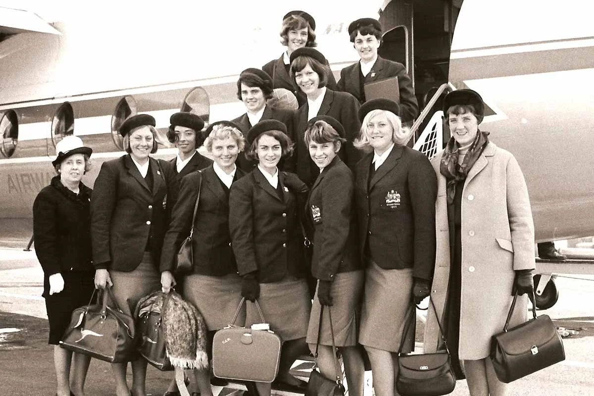 A group of women in vintage flight uniforms standing in front of an airplane, holding bags and wearing hats, smiling for the camera.