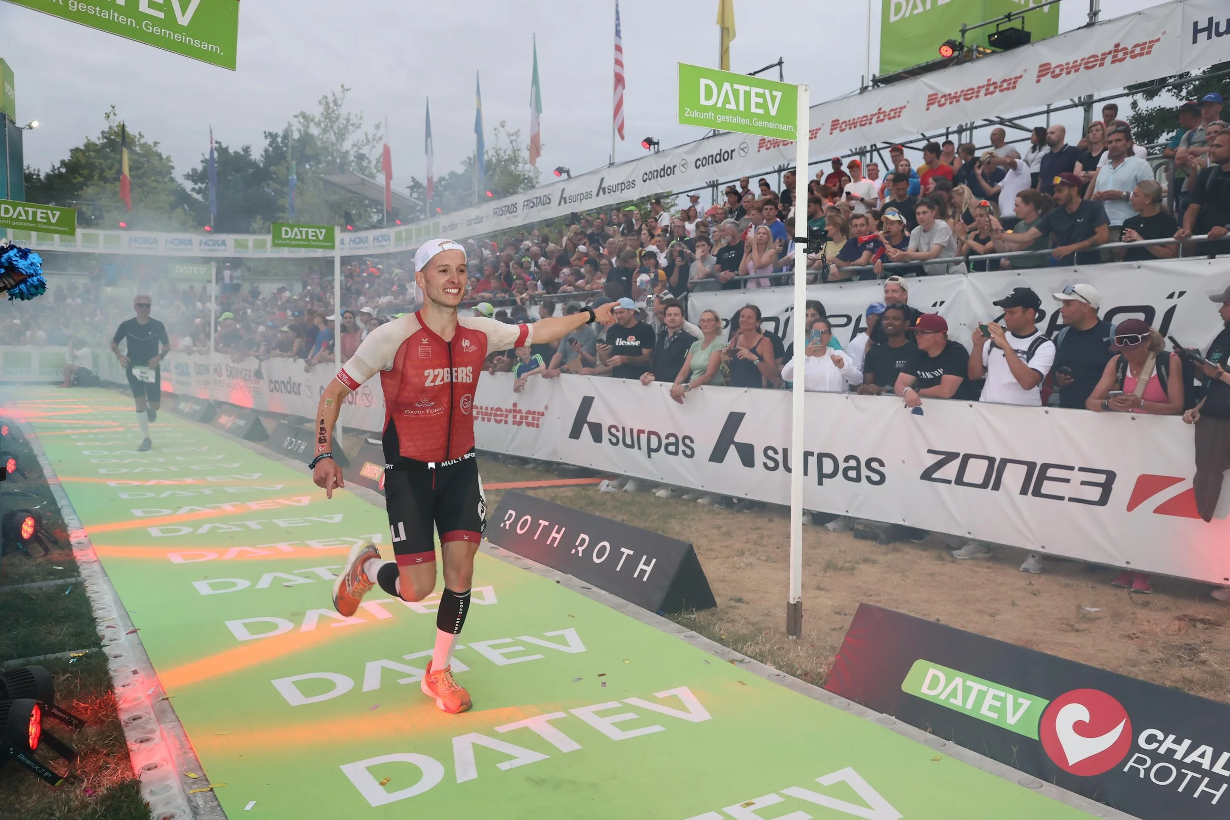 A male athlete running and smiling near the finish line at a race event called DATEV Challenge Roth, with a cheering crowd in the background.