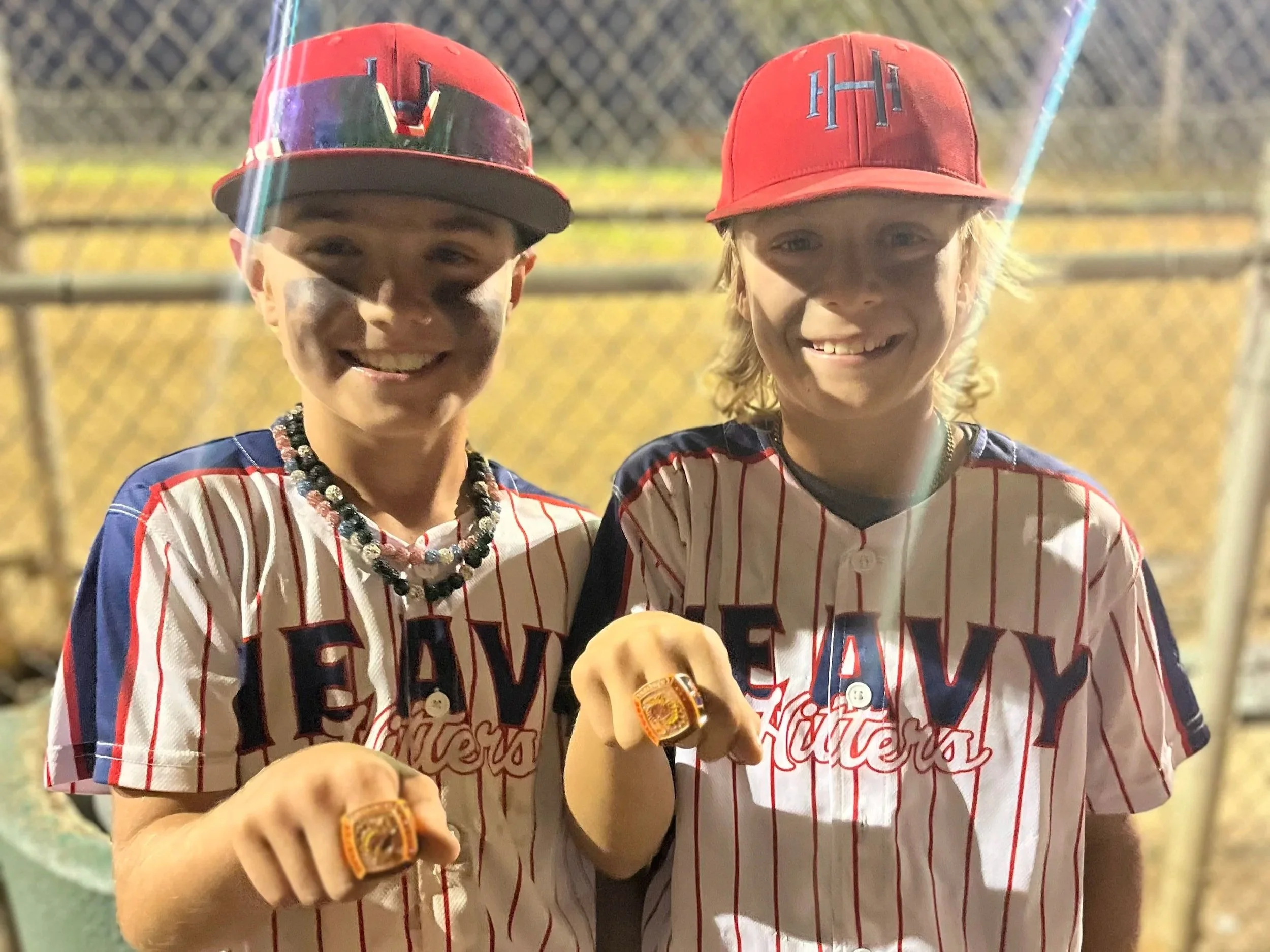 Two young boys wearing baseball jerseys and red caps, each displaying a championship ring, standing outdoors near a chain-link fence at night.