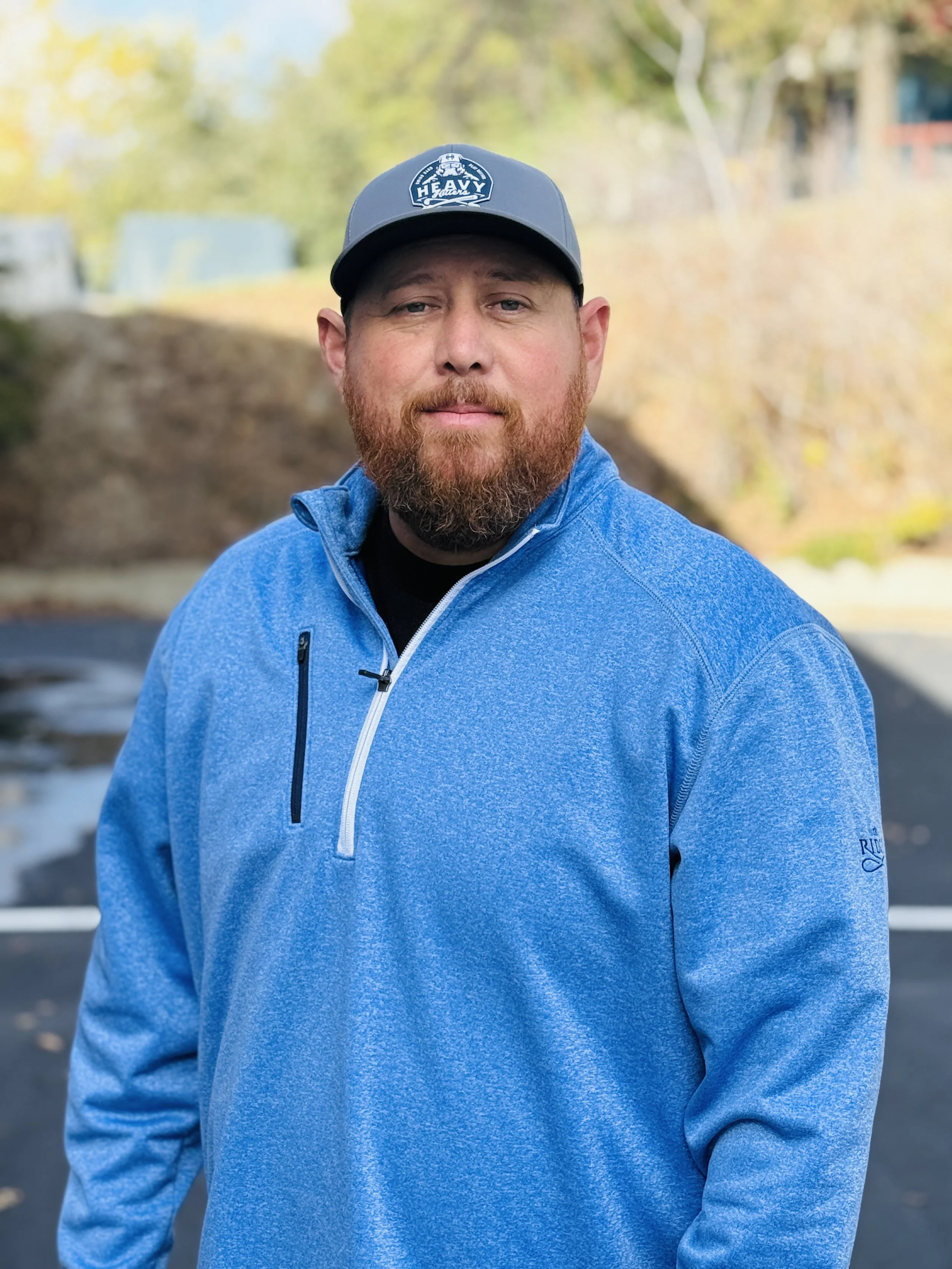 A man with a beard and mustache wearing a blue quarter-zip jacket and a gray baseball cap standing outdoors on a paved surface with trees in the background.